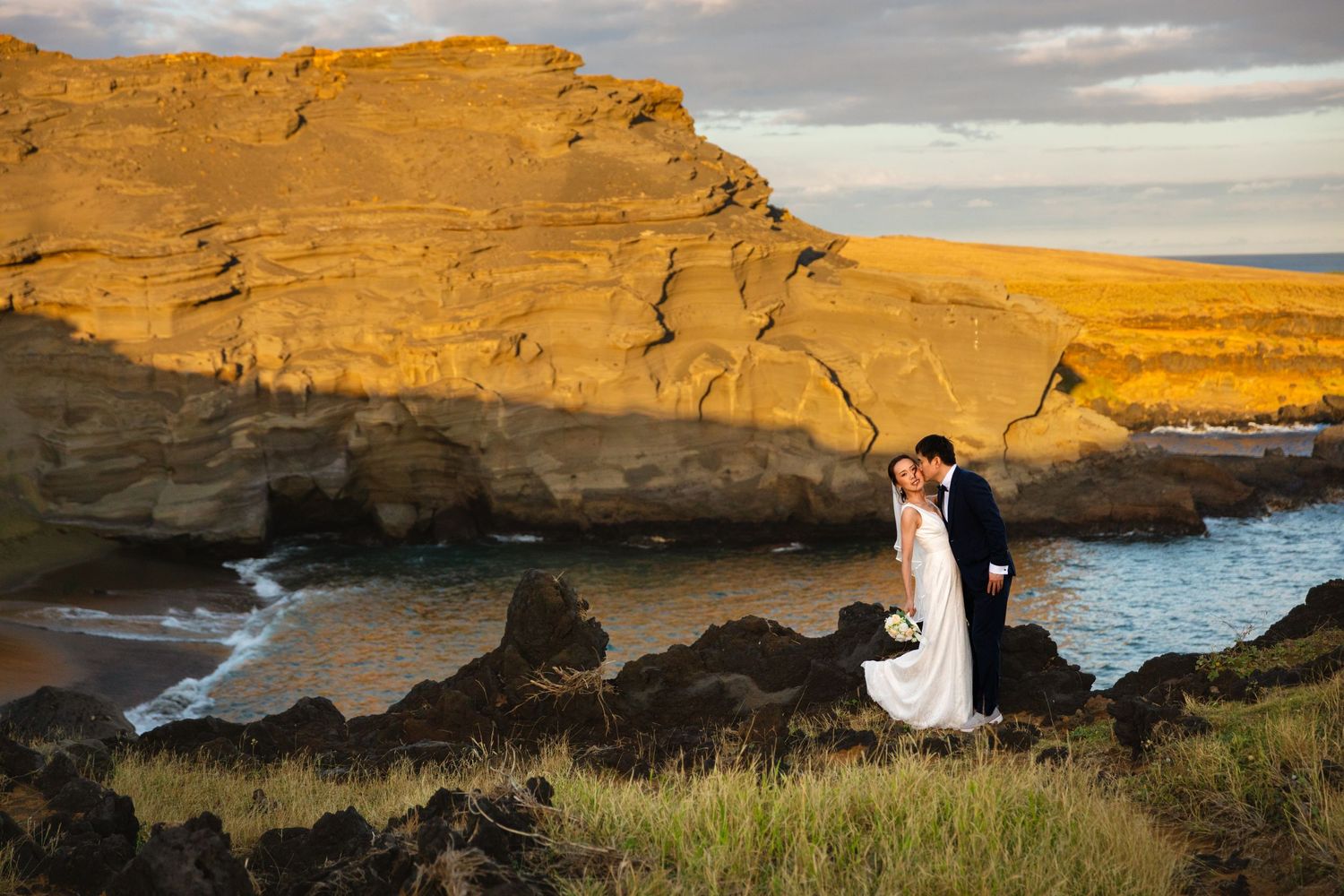 Bride and groom kissing at golden hour beside dramatic coastal cliffs and turquoise ocean waters in Hawaii.