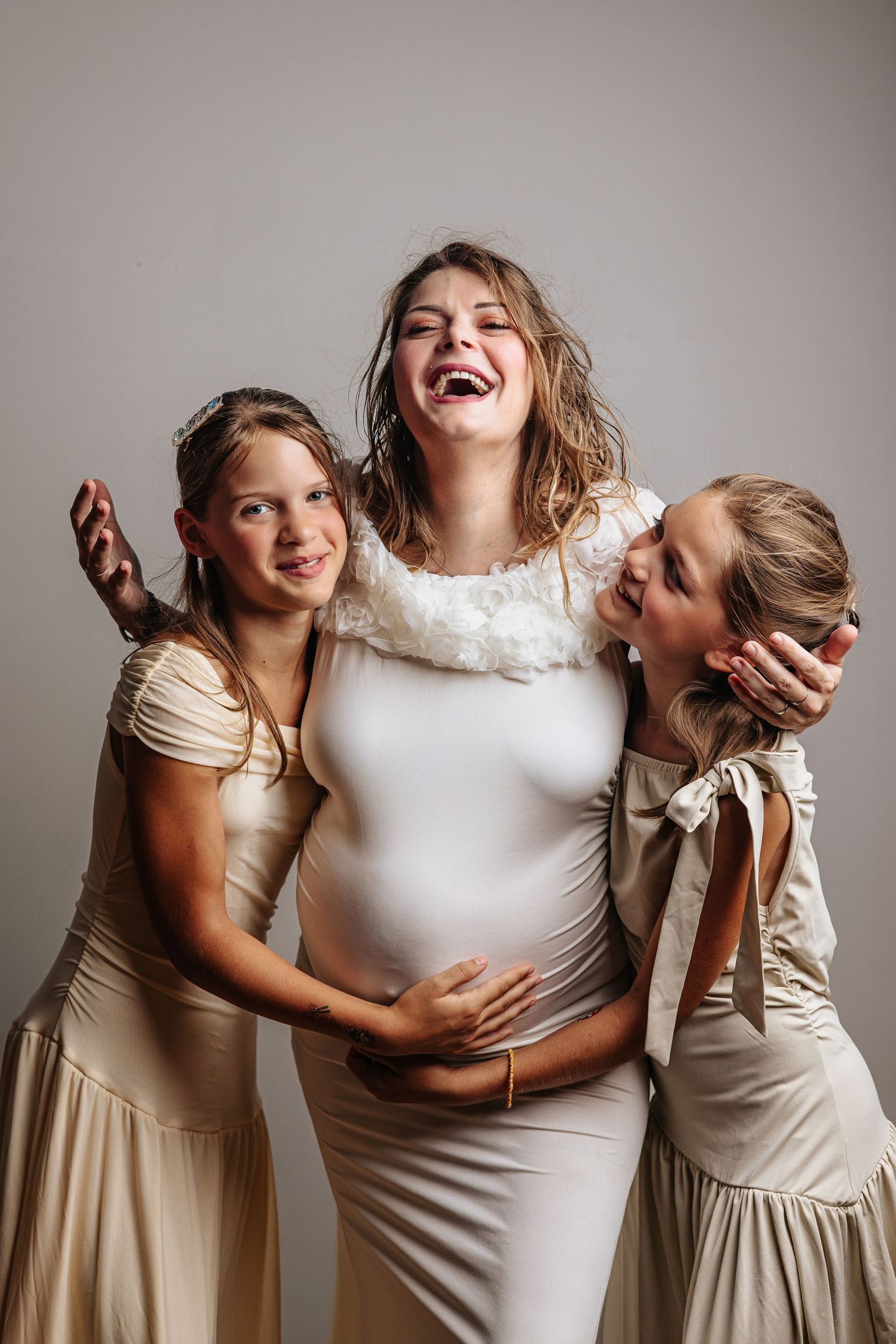 Pregnant woman in white dress laughing joyfully with two young girls in beige dresses hugging her baby bump.