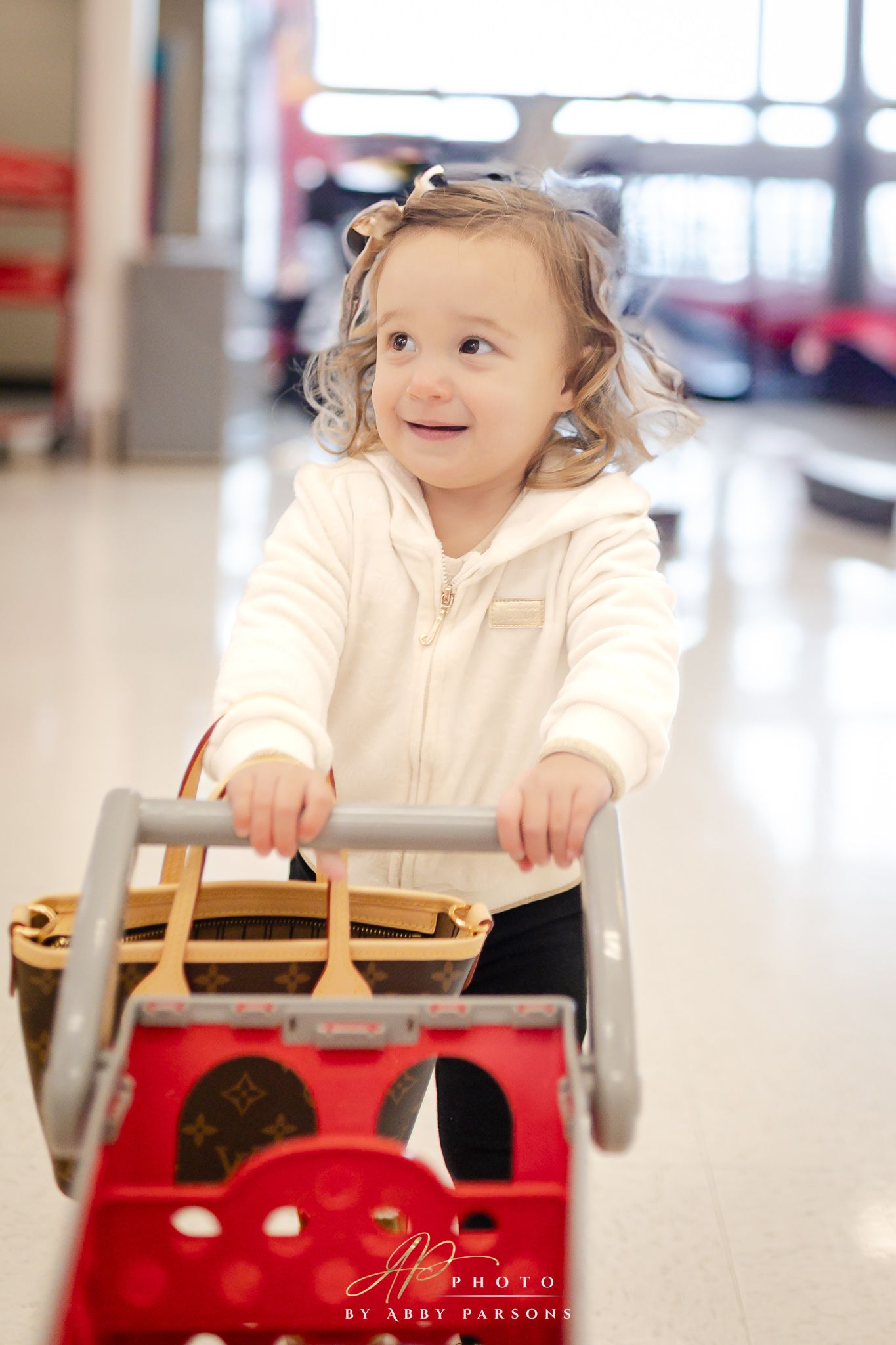 A toddler in a cream-colored hoodie pushes a red toy shopping cart in a bright indoor space.