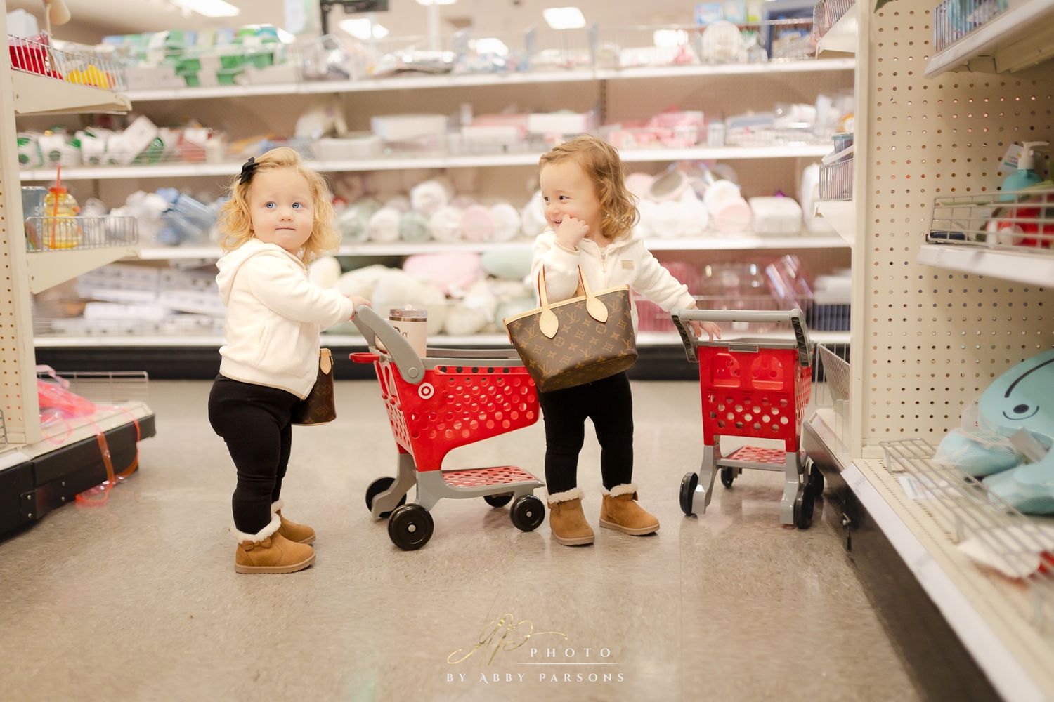 Two toddlers in winter boots push tiny red shopping carts down a store aisle filled with retail shelves.