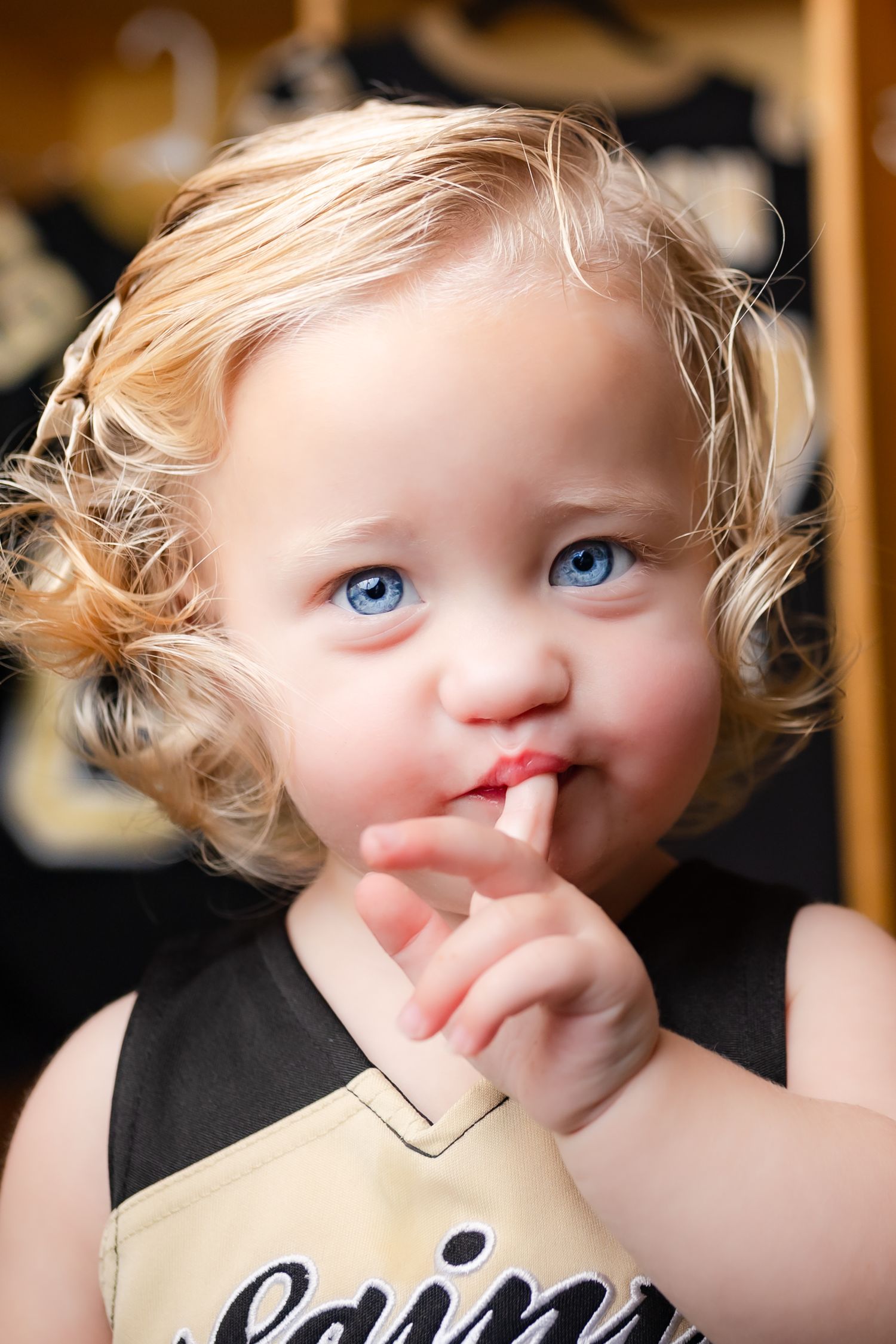 A small child with blonde curly hair and bright blue eyes holds a finger to their lips in a playful gesture.