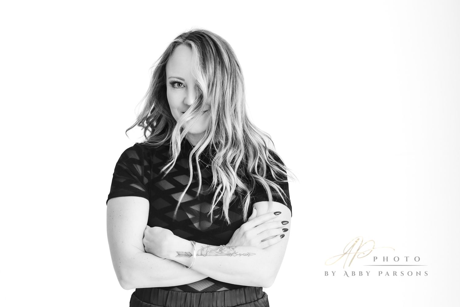 Black and white studio portrait of a person with long wavy blonde hair wearing dark clothing and standing with arms crossed.
