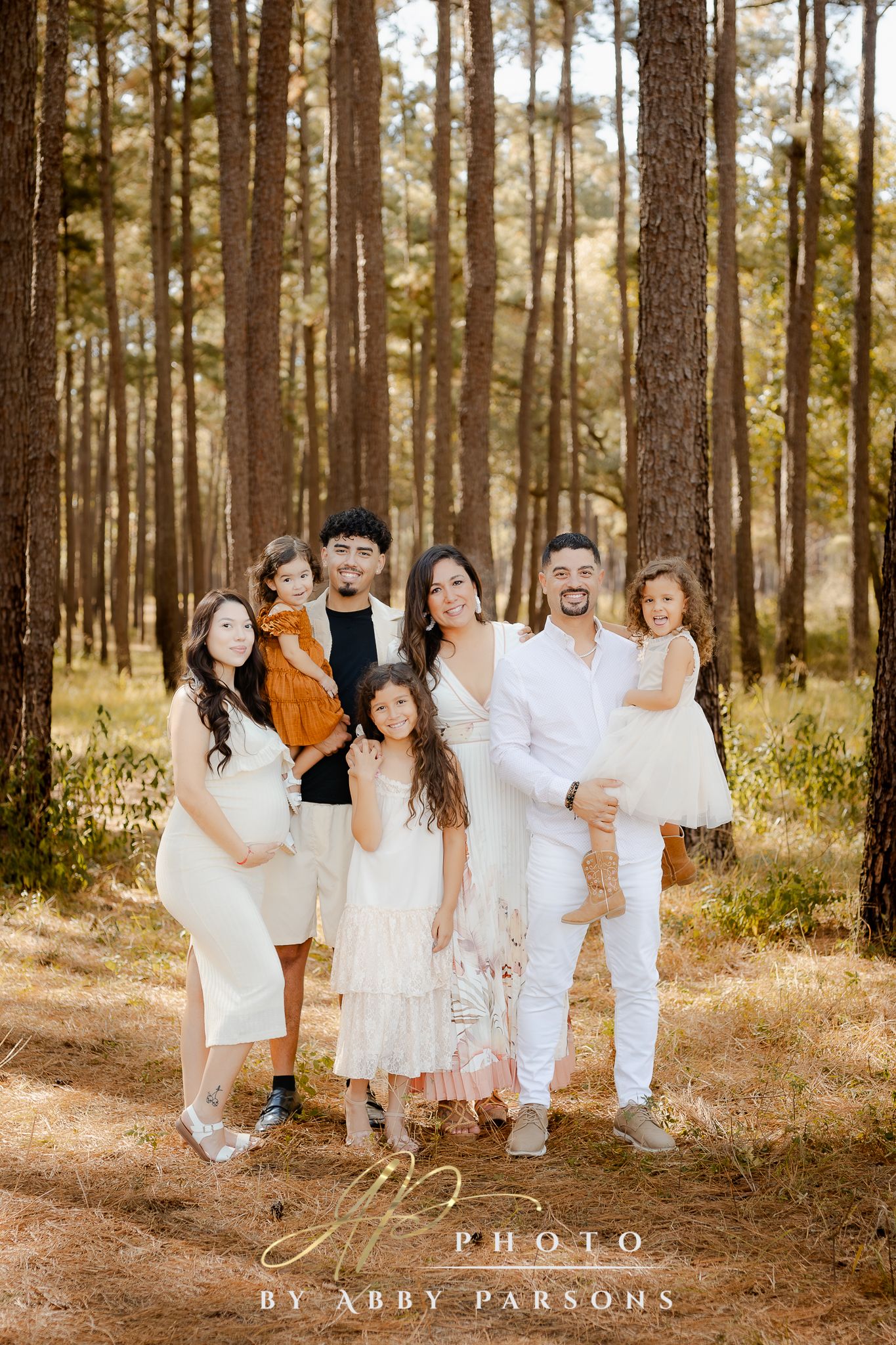 A group in coordinated white and neutral outfits poses together in a sunlit pine forest setting.