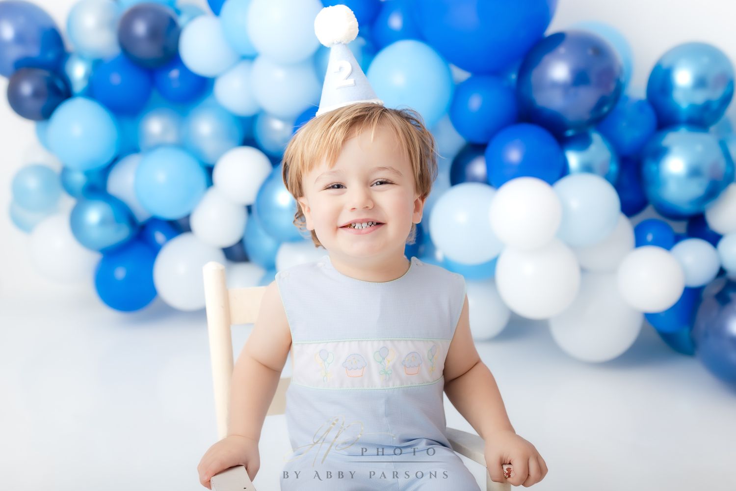 A young child smiles while sitting in front of a festive backdrop of blue and white balloons.