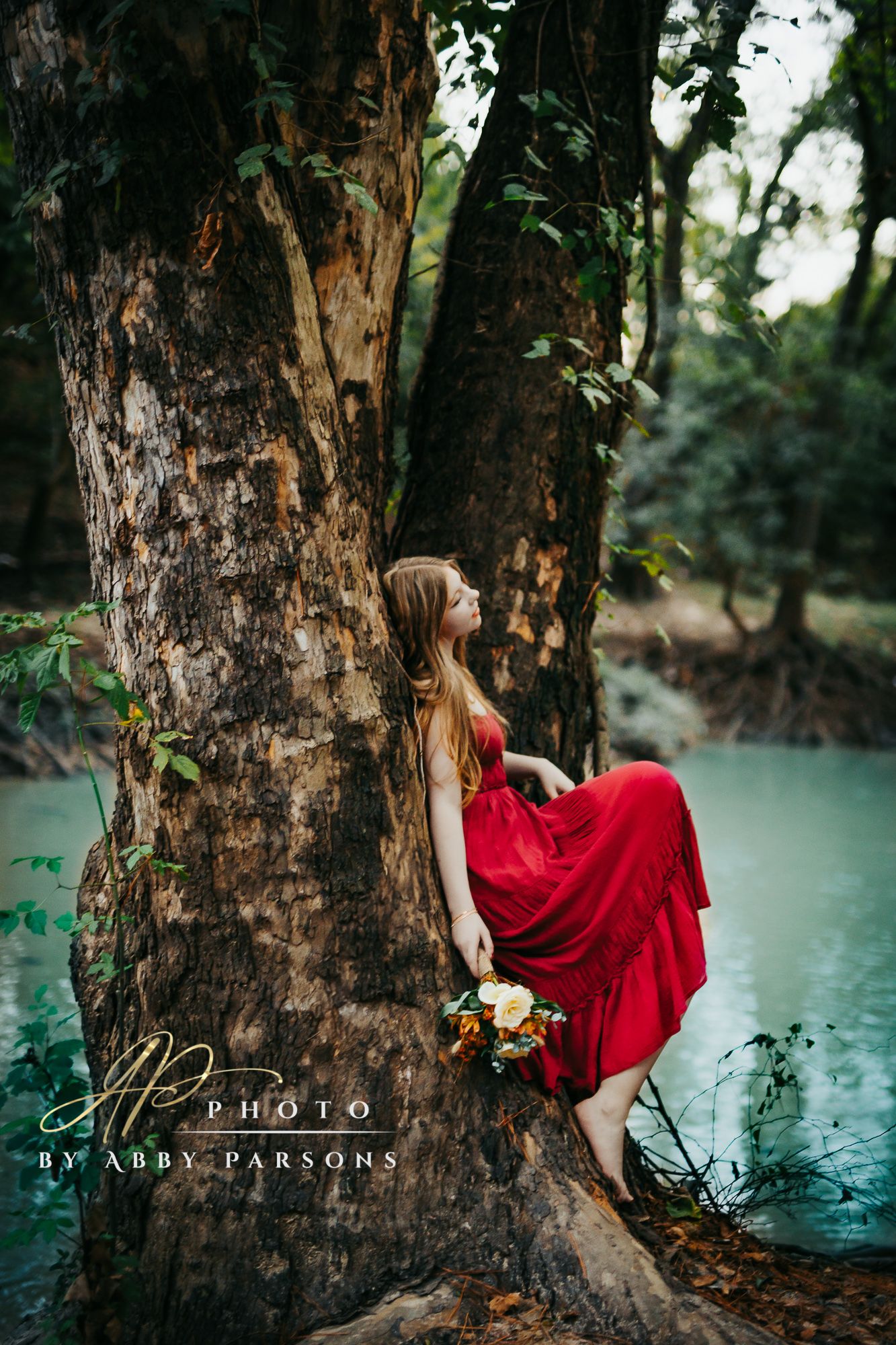 A figure in a flowing red dress leans against a large tree trunk beside a tranquil turquoise pond in a moody forest setting.
