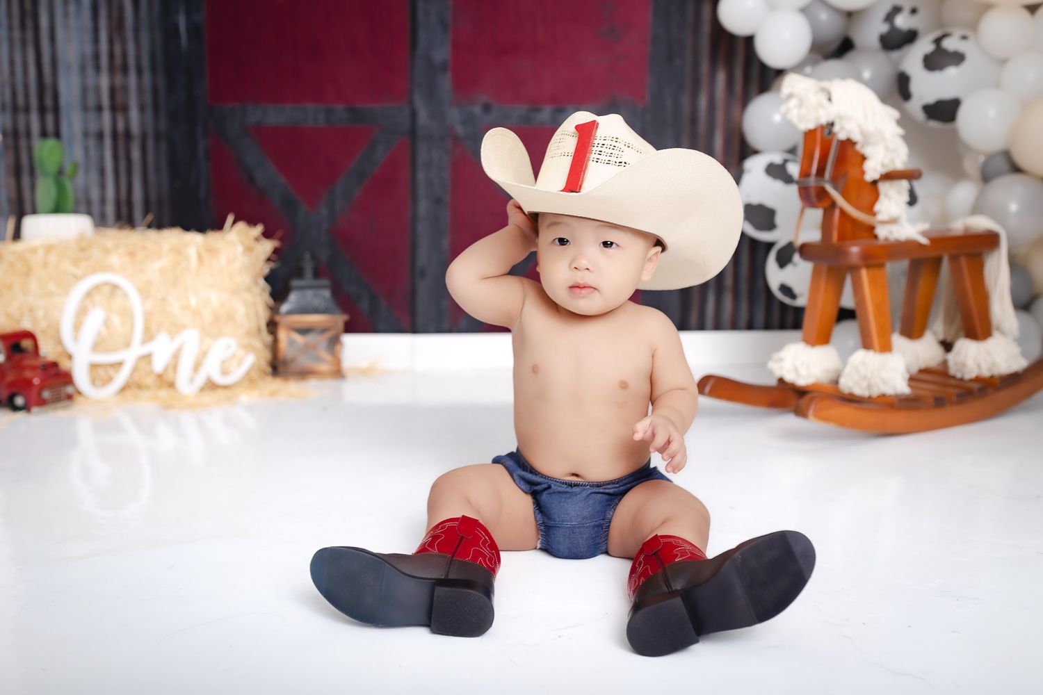Baby in cowboy hat and boots sits on white floor with rustic barn door and rocking horse in background.