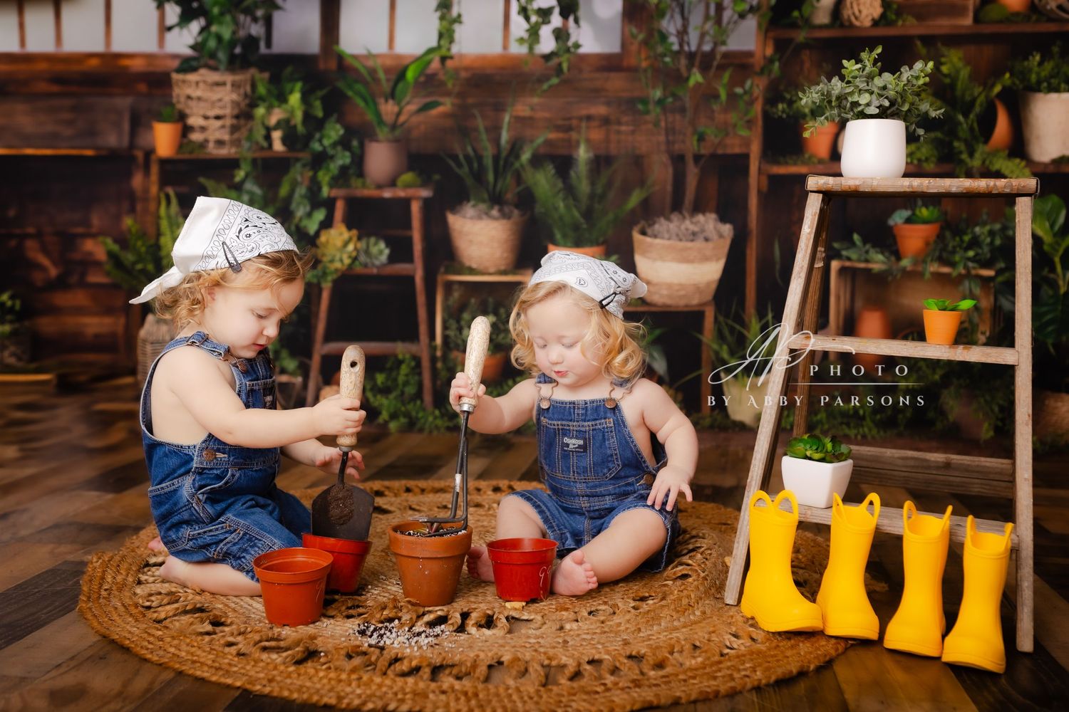 Two young children in denim overalls play with gardening tools on a round woven mat surrounded by potted plants and yellow boots.