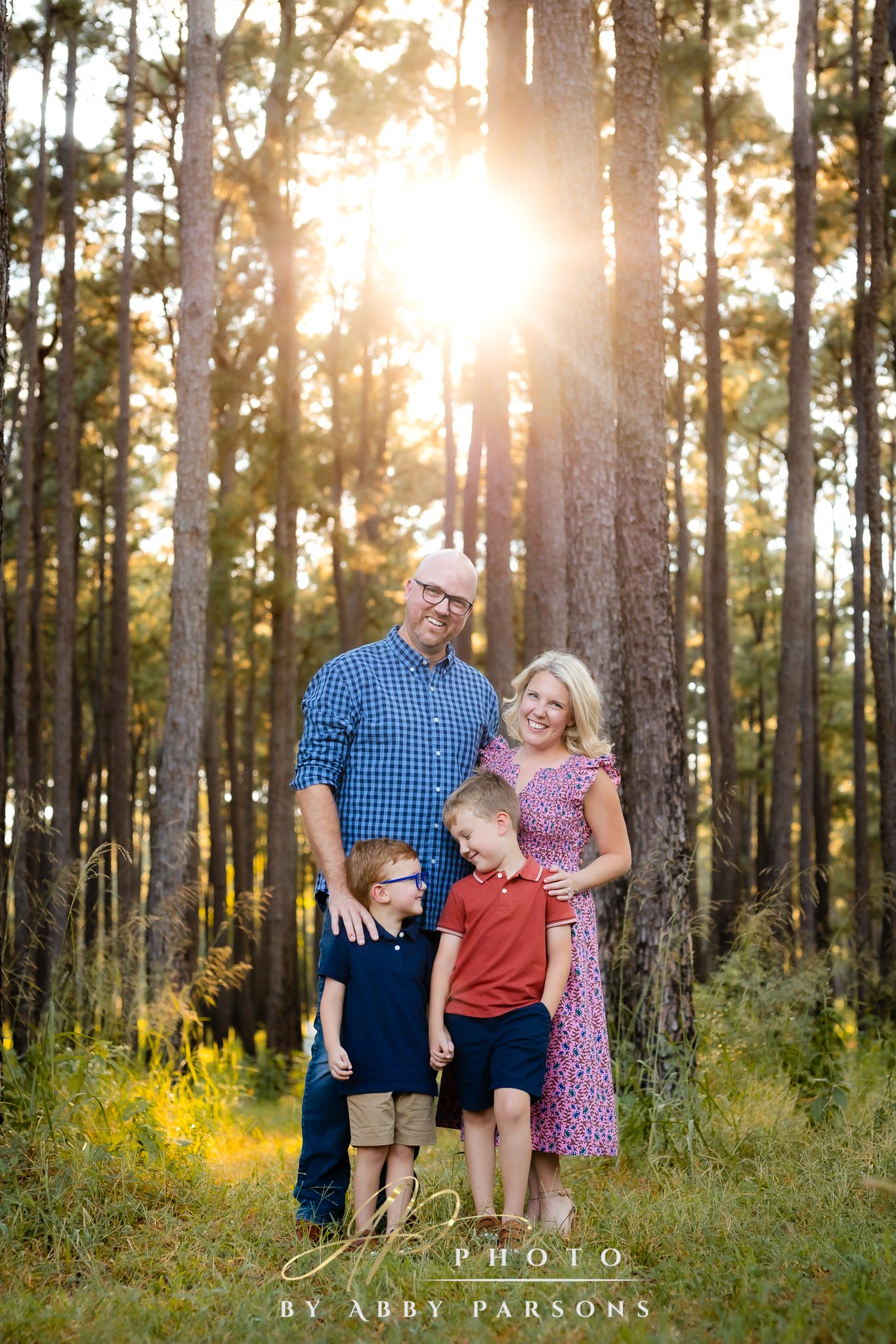A family poses together in a sunlit forest with pine trees and yellow wildflowers at golden hour.
