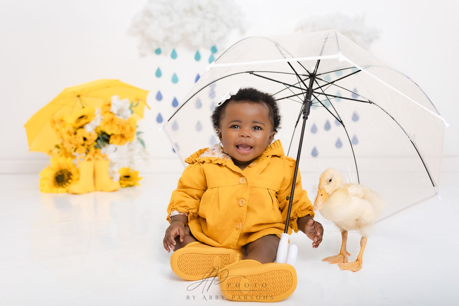 A child in bright yellow rain gear sits with a clear umbrella and rubber duck next to yellow sunflowers against a white backdrop.