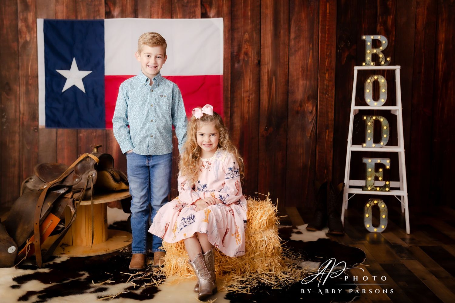 Two young children pose in front of a Texas flag with rustic barn wood background and western decor.
