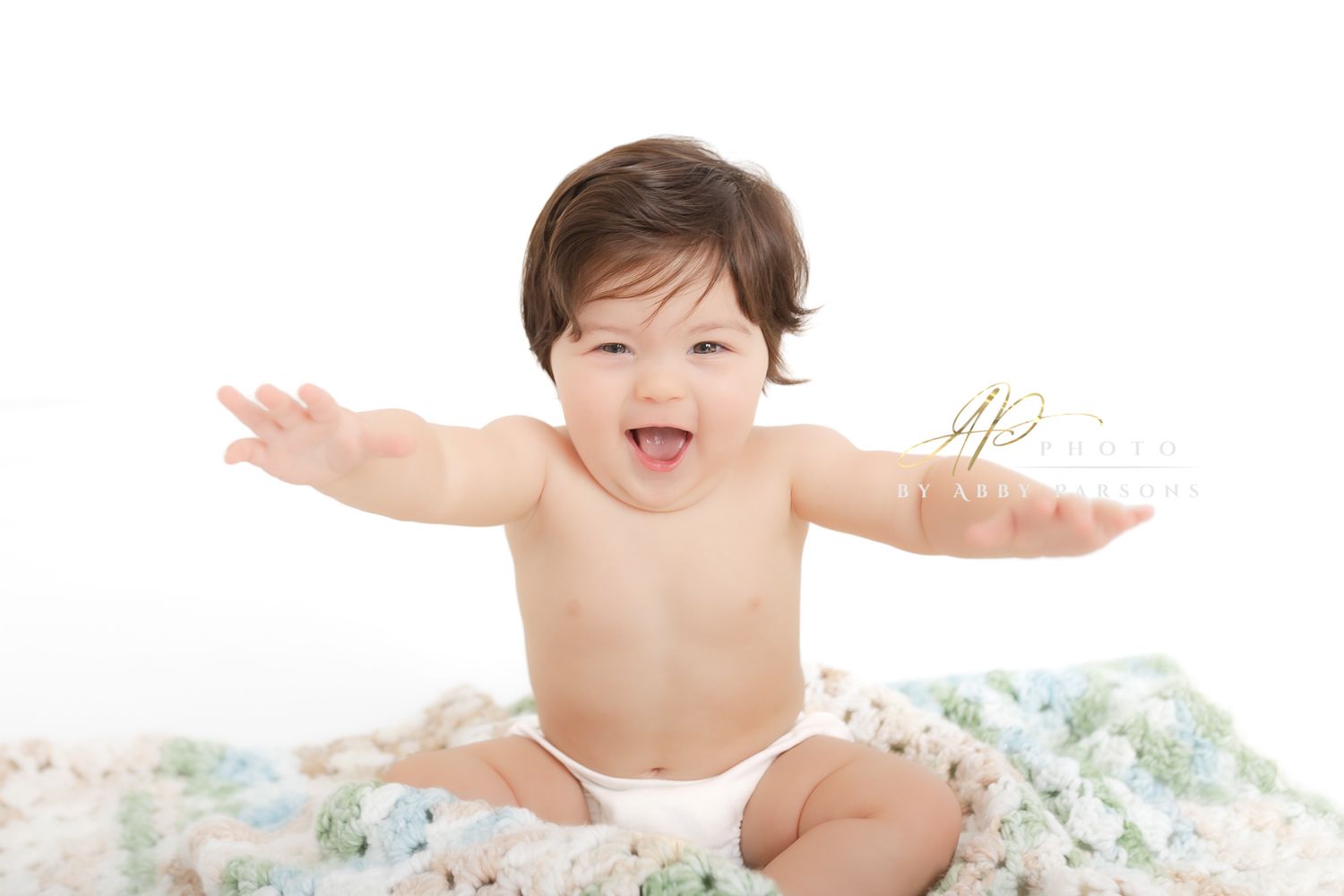 Baby wearing a white diaper smiles with arms outstretched while sitting on a floral blanket.