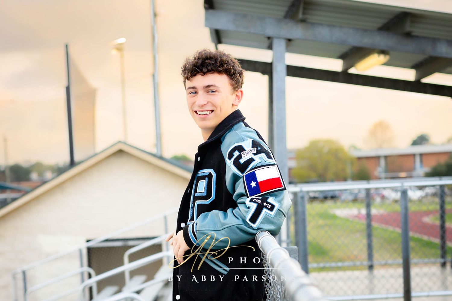 High school athlete in varsity letterman jacket with team number 24 poses at sports field during sunset.