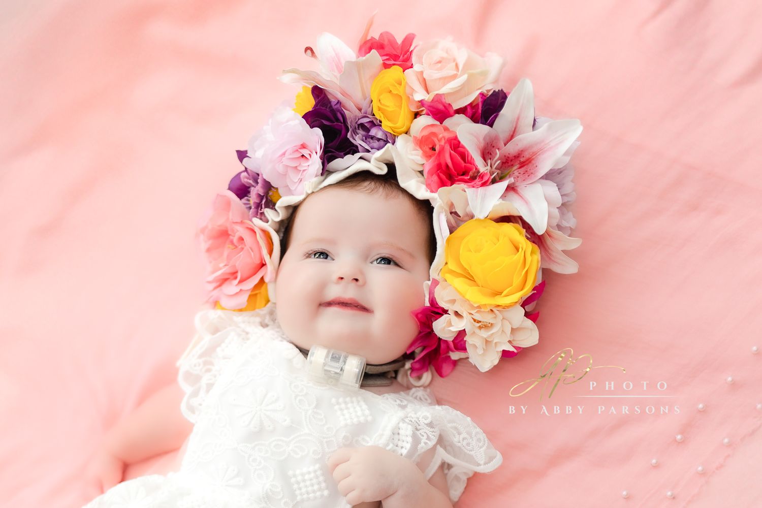Baby smiling while wearing a white dress and colorful floral crown headpiece on pink background.