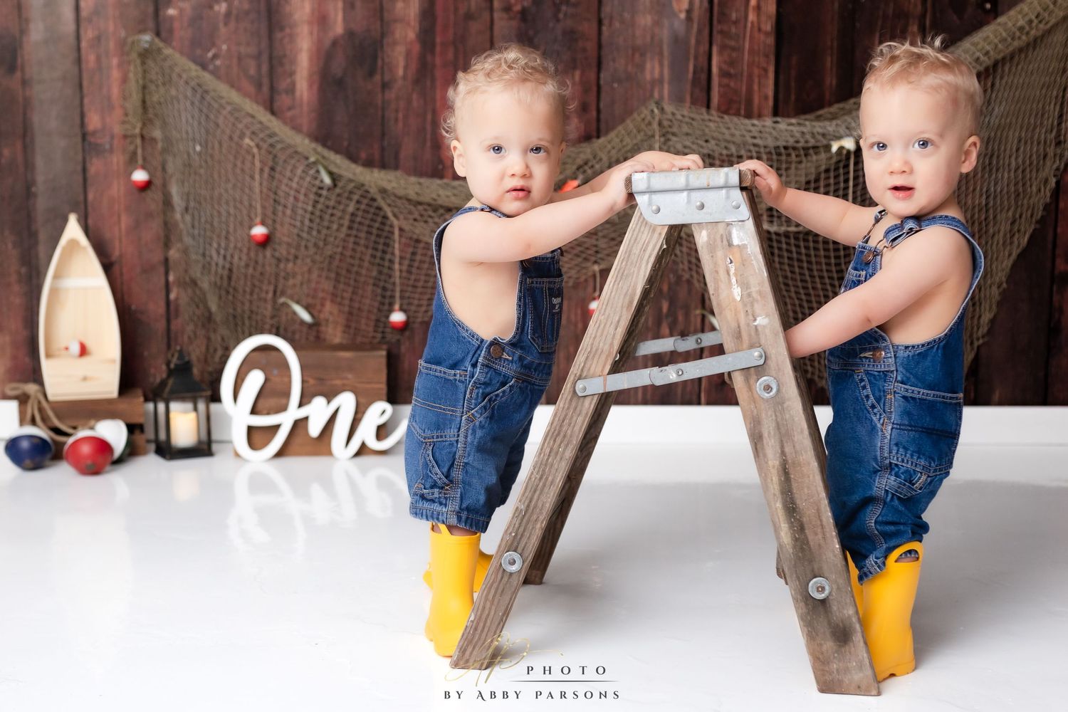 Two toddlers in matching denim overalls and yellow boots stand at a wooden ladder with a rustic backdrop.