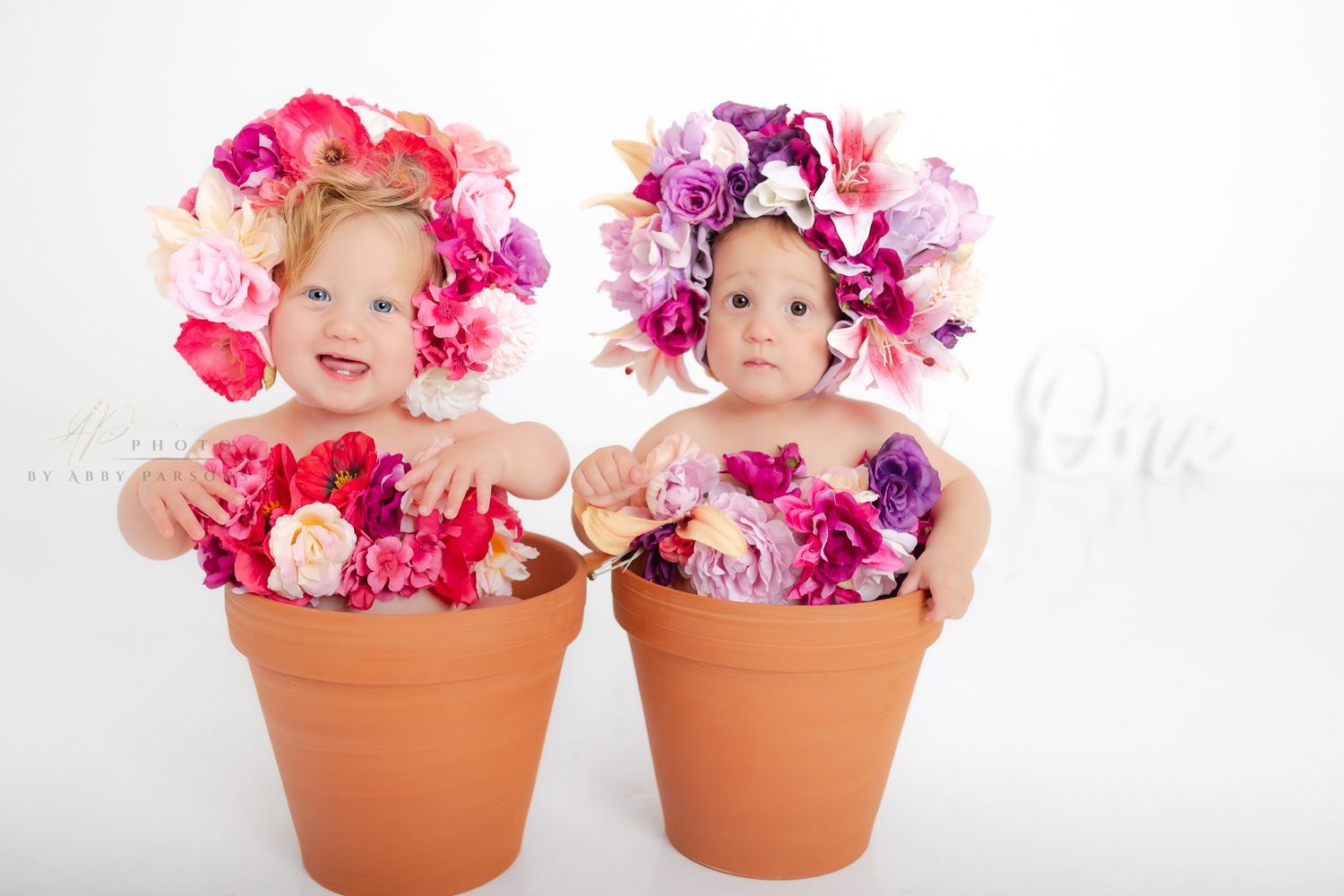 Two toddlers dressed as flowers sit in terracotta pots with colorful pink and purple floral decorations.