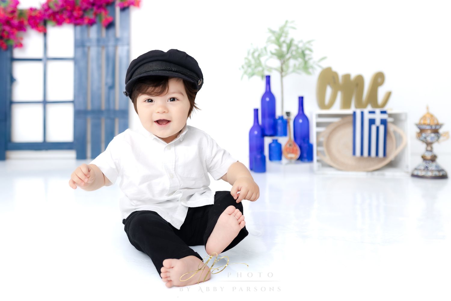 A baby wearing a white shirt and black hat sits on a white floor with blue glass bottles in the background.