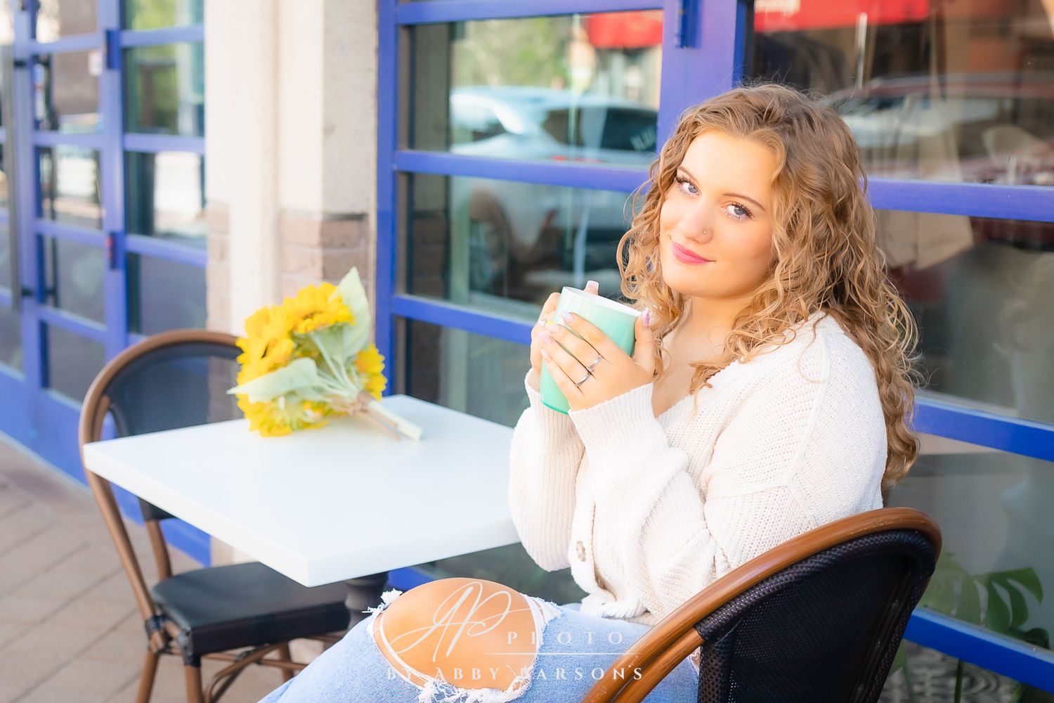 Someone enjoys coffee while relaxing at an outdoor cafe table with yellow flowers and blue storefront in background.