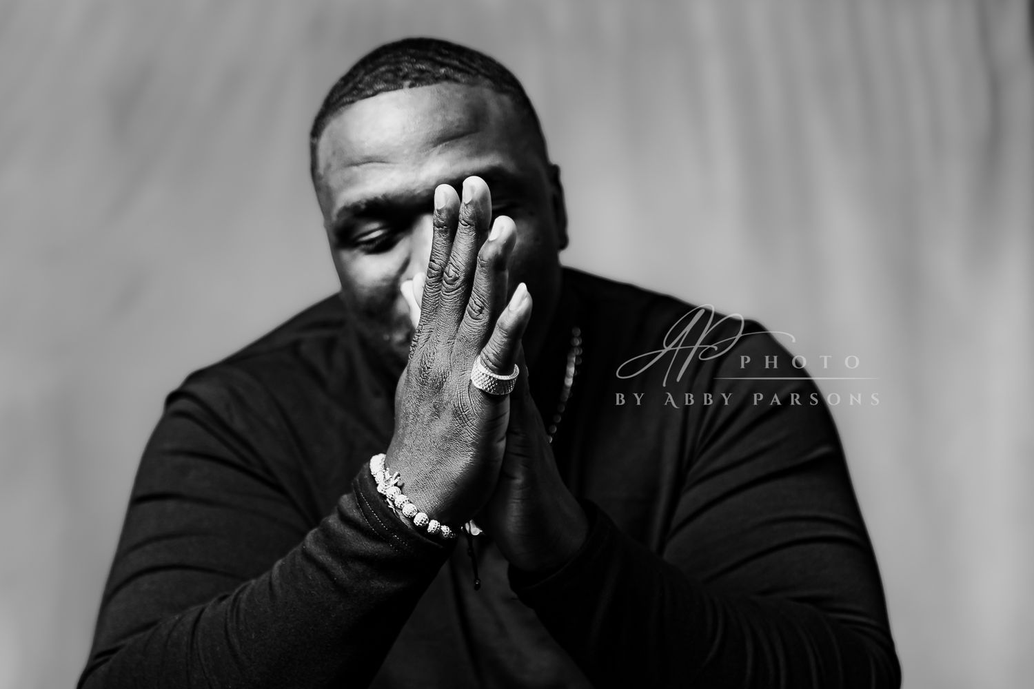 Black and white portrait shows person in dark sweater covering face with one hand while wearing silver bracelet.