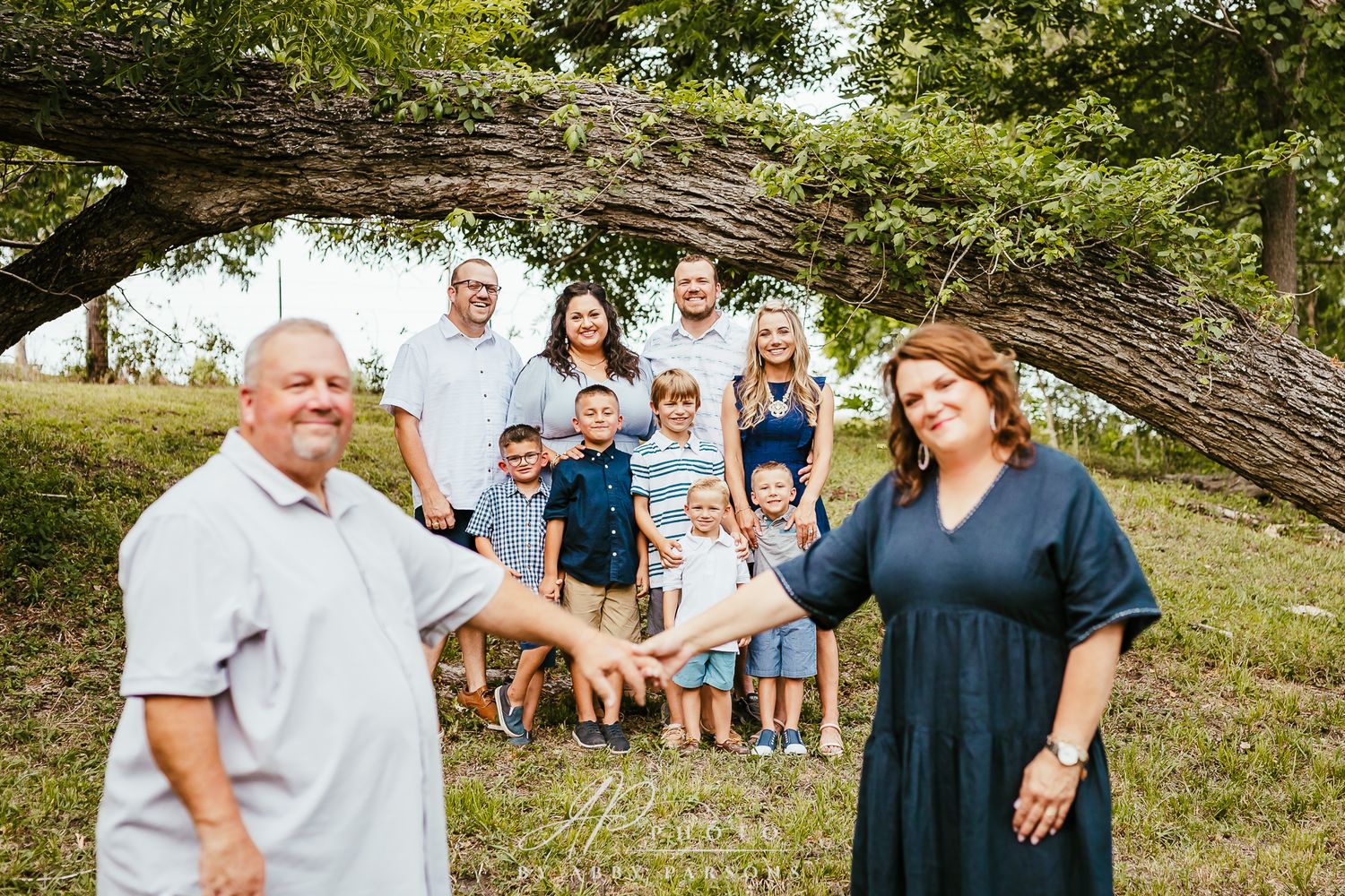 A large family group gathers under a leaning tree branch on a grassy hill on a sunny day.