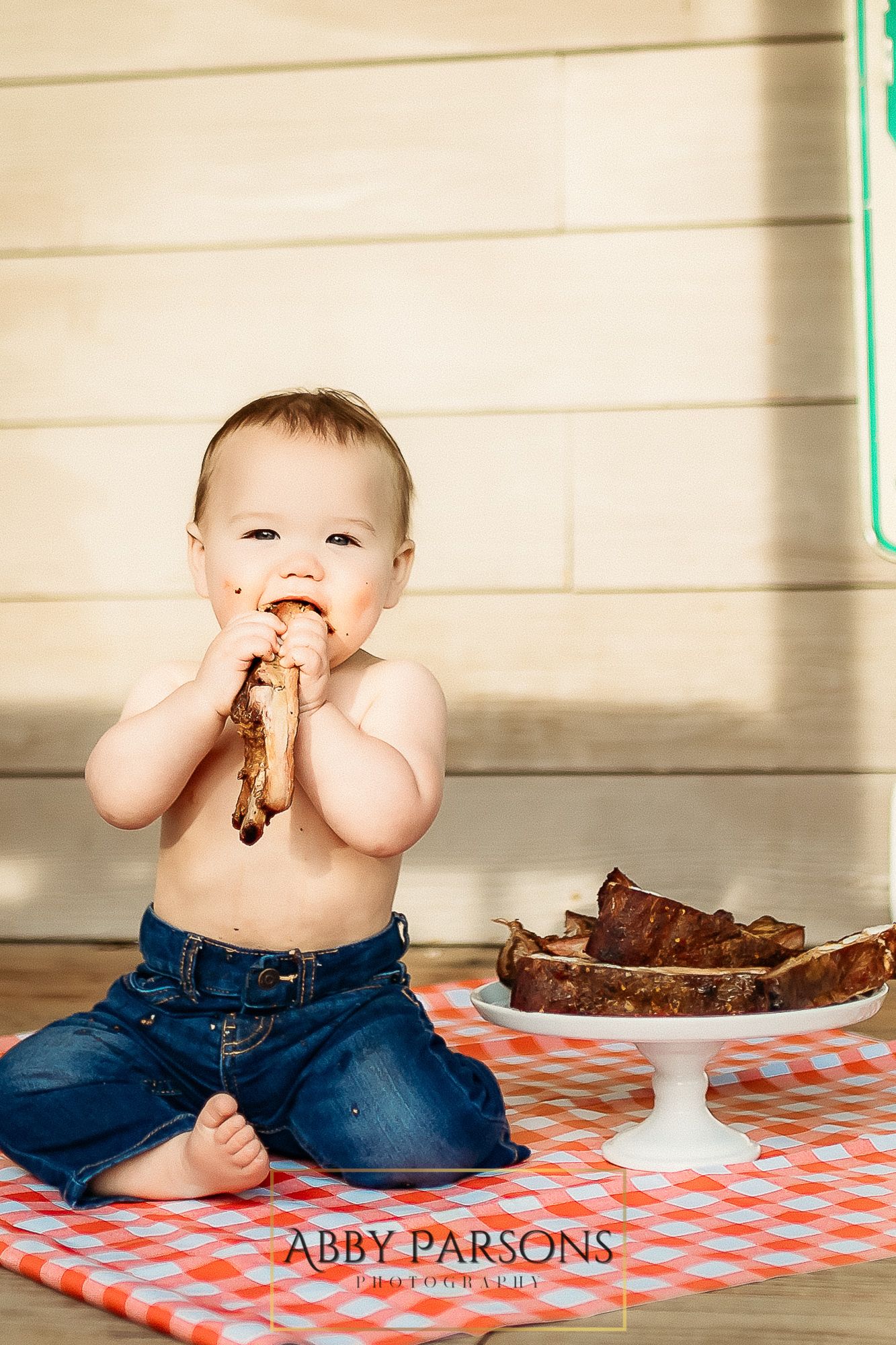 Small child in jeans sits on red checkered blanket enjoying chocolate cake slice from white cake stand.