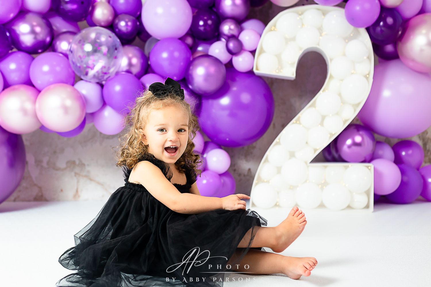 Toddler in black tulle dress poses with large number 2 against purple balloon backdrop.