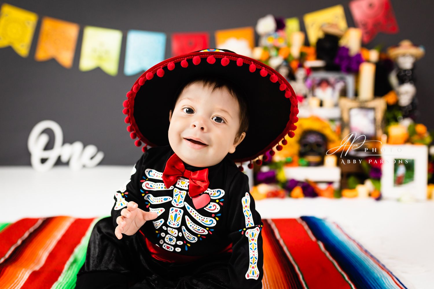 A small child in a skeleton costume with red sombrero smiles at a festive party with colorful decorations.