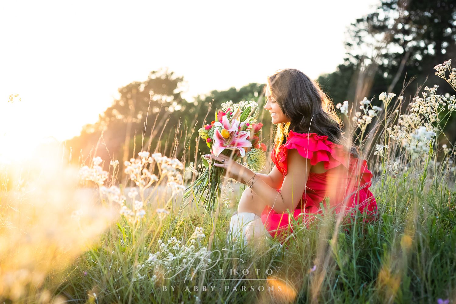Woman in bright red dress sitting among wildflowers during golden sunset in meadow field.