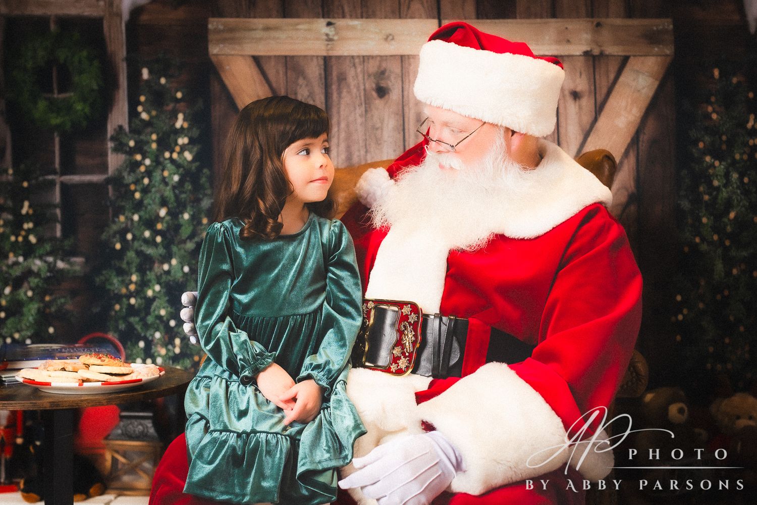 A Christmas scene with Santa in traditional red suit seated with a child in a teal velvet dress.