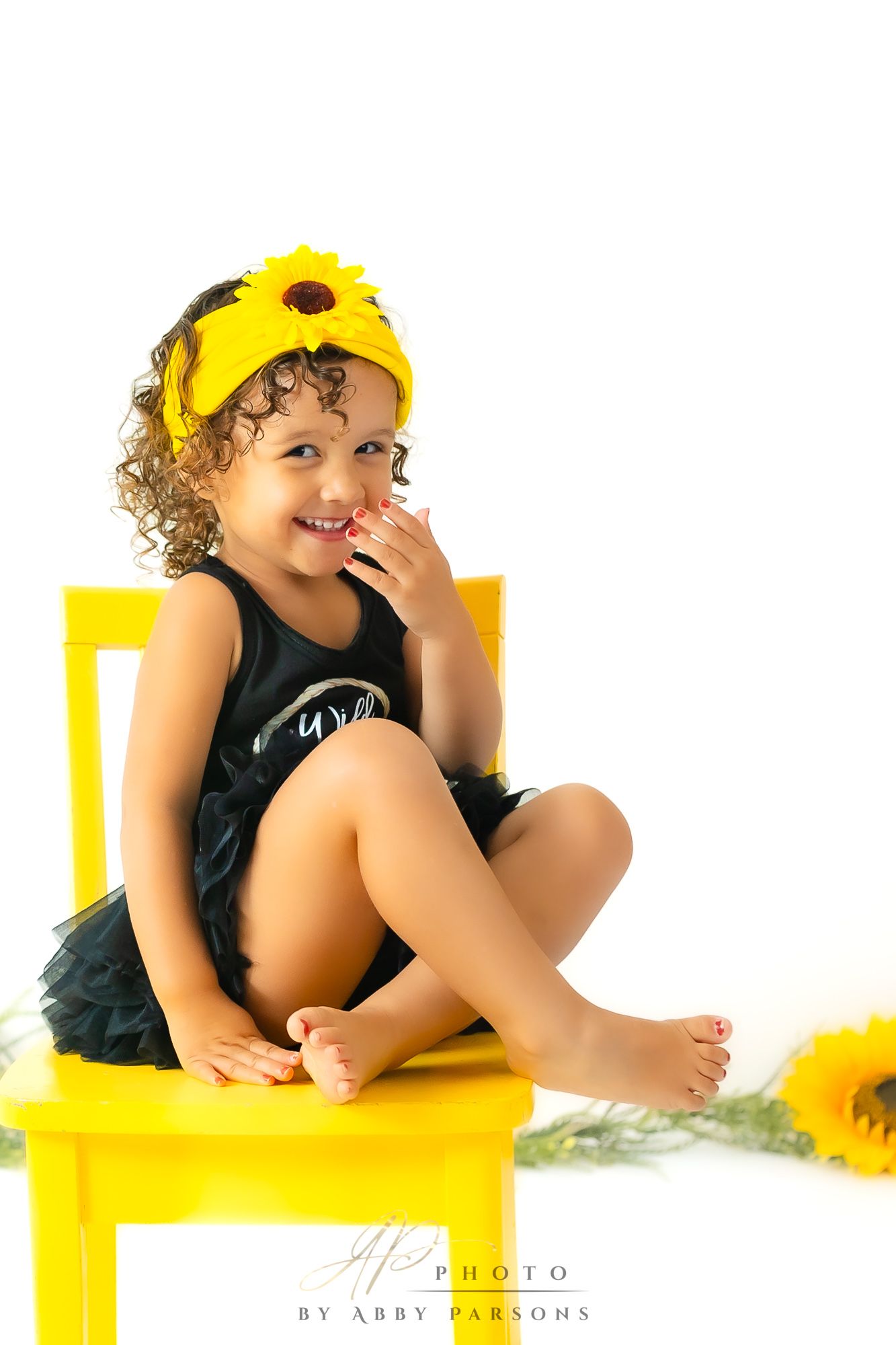 A small child wearing a yellow sunflower headband and black outfit sits playfully on a bright yellow chair.