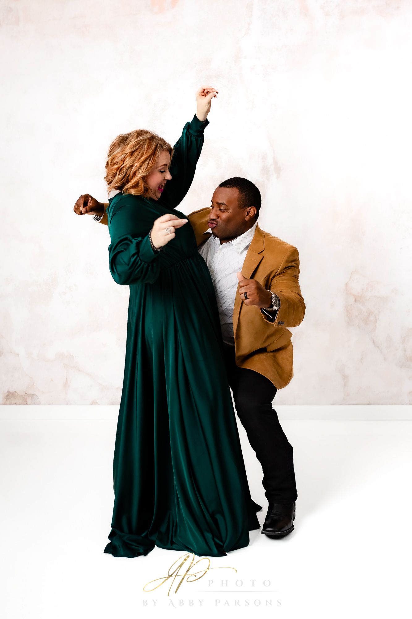 Two people in formal attire dancing joyfully against a white backdrop, one in emerald green and one in tan.