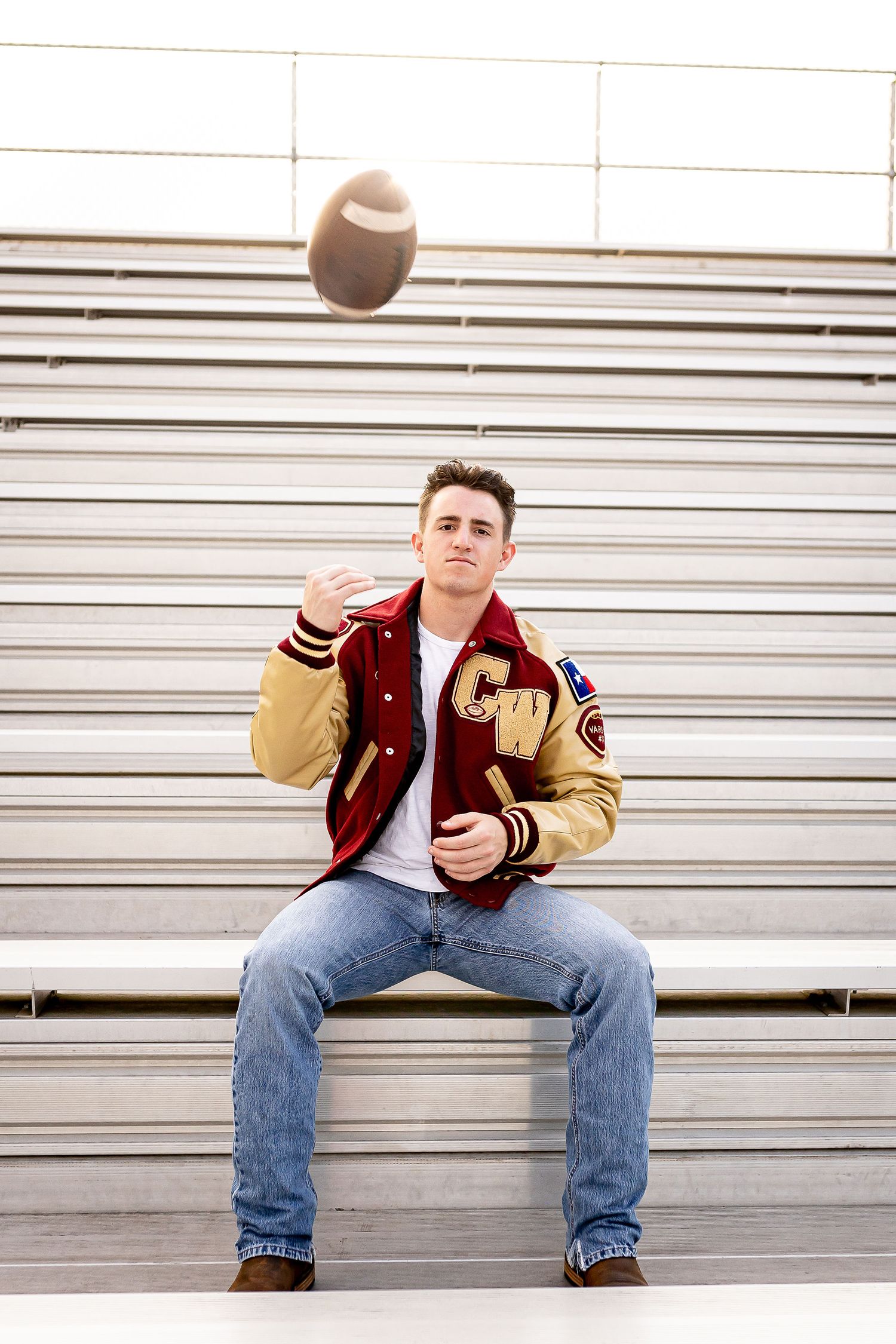 An athlete in a varsity letterman jacket sits on bleachers while tossing a football in the air.