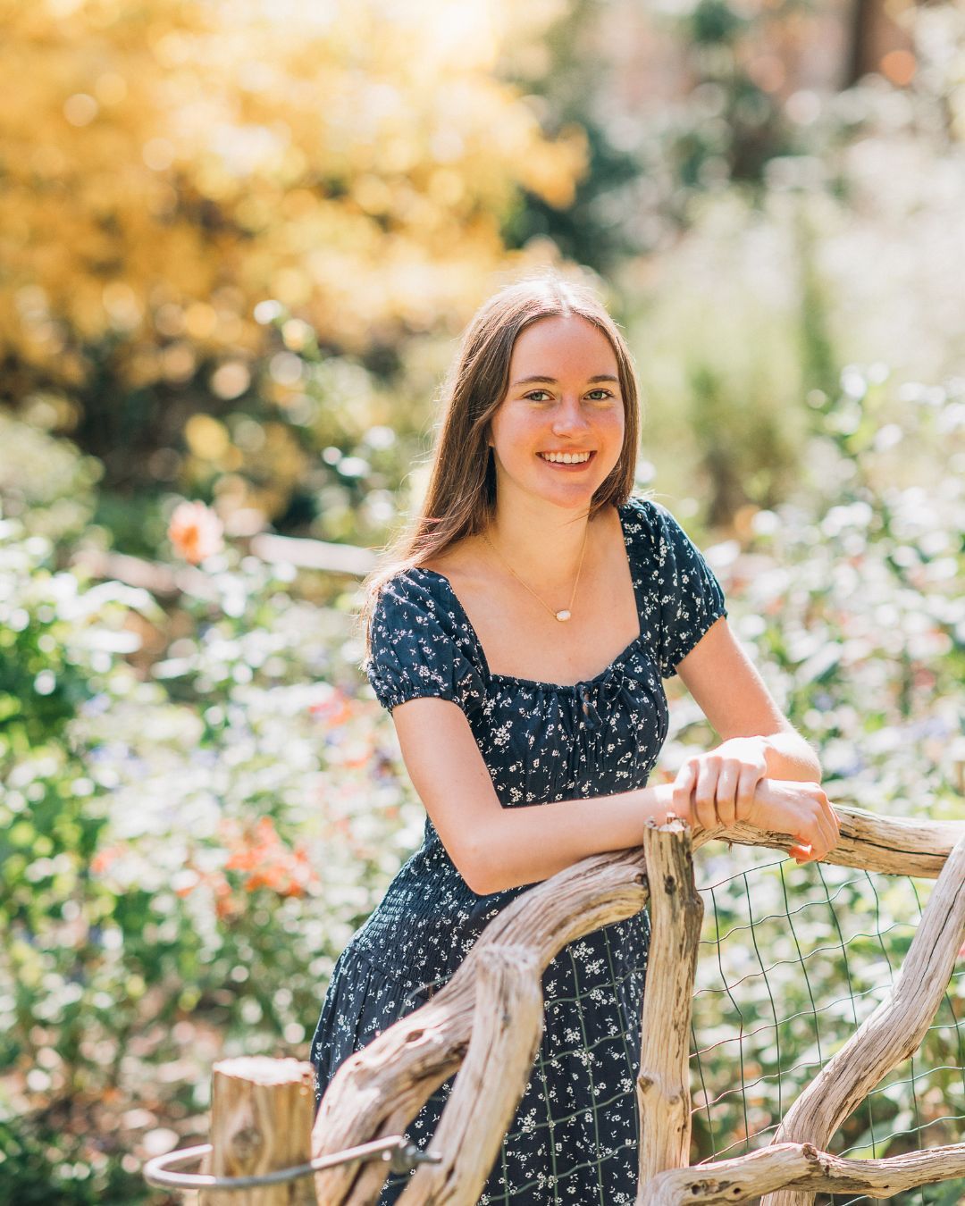 Portrait in floral garden with person in navy dress seated on rustic wooden chair.