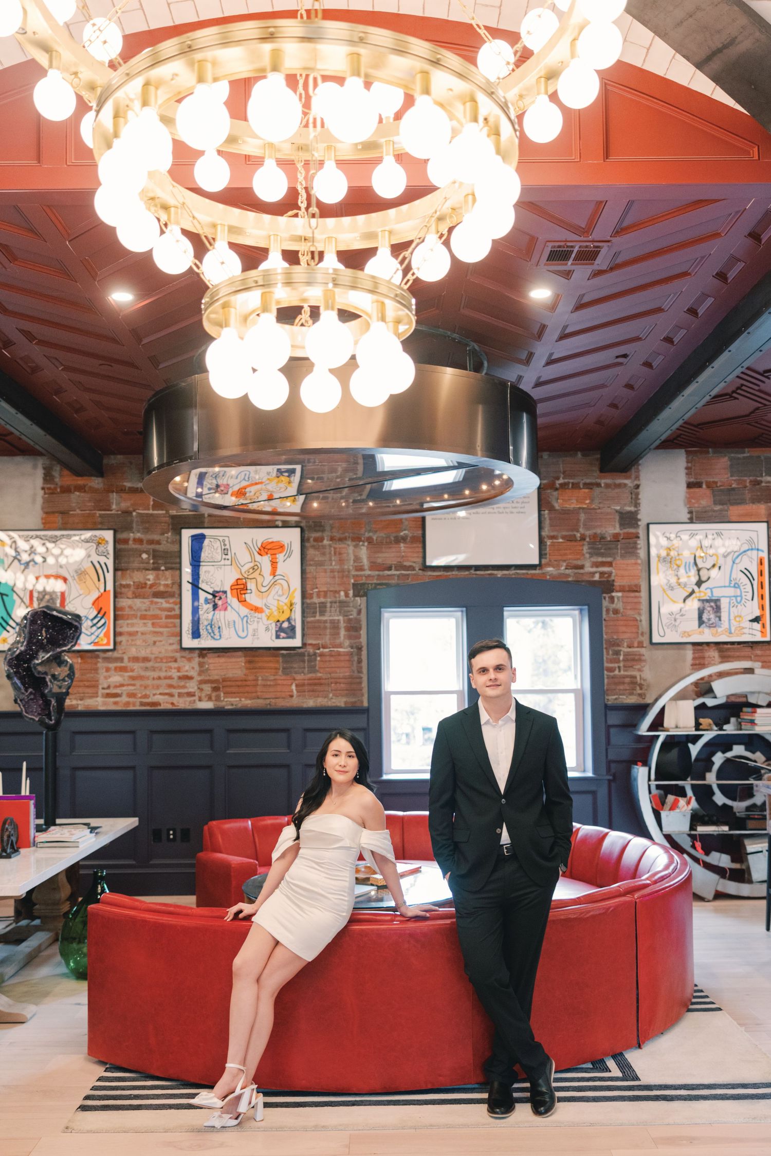 Couple posing in a stylish restaurant with red sofa and decorative chandelier for engagement photos.