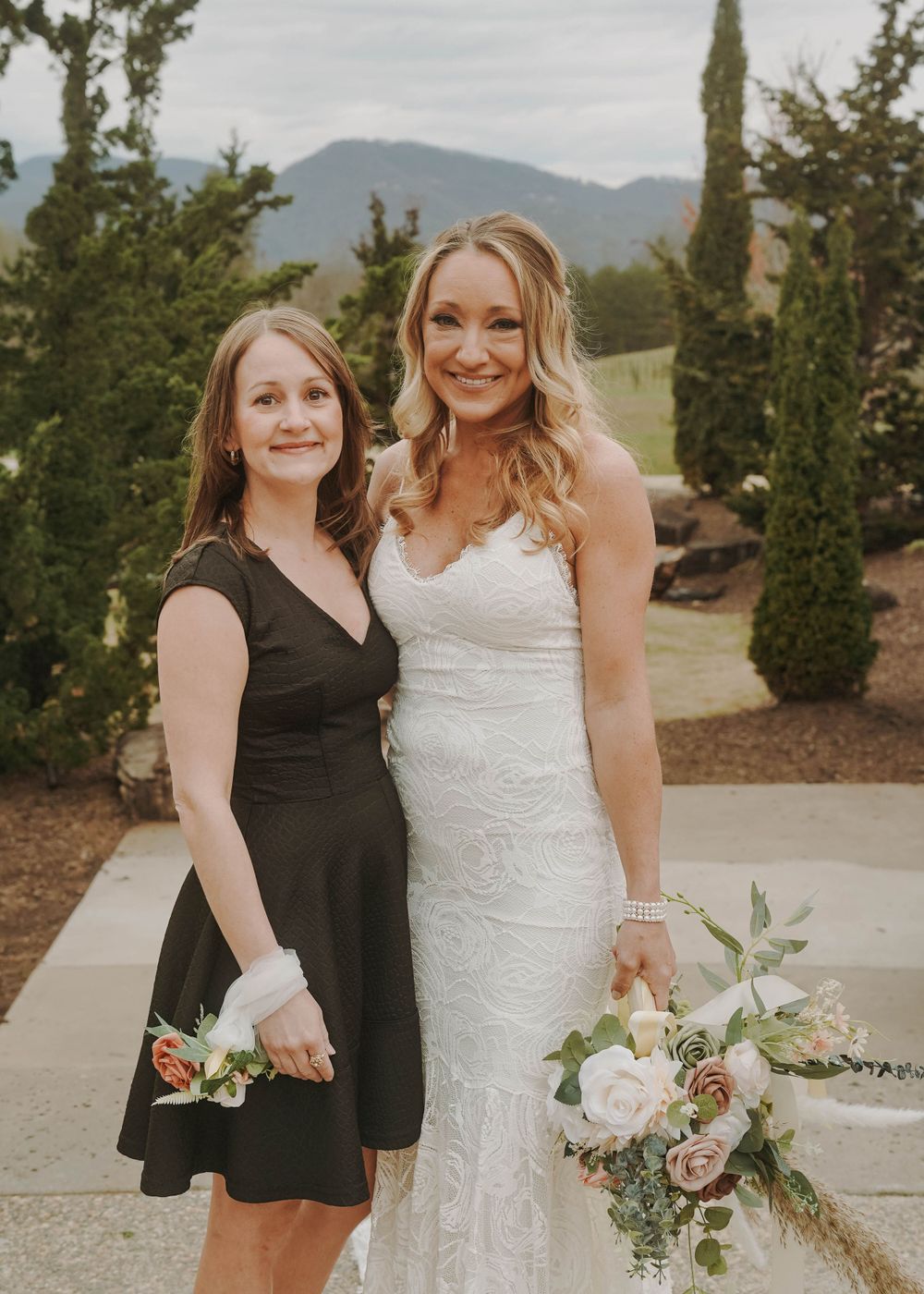 Two people posing with bouquets against mountain landscape at wedding event.