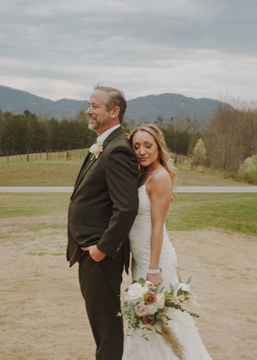 Couple in wedding attire pose back-to-back with mountain landscape in background.