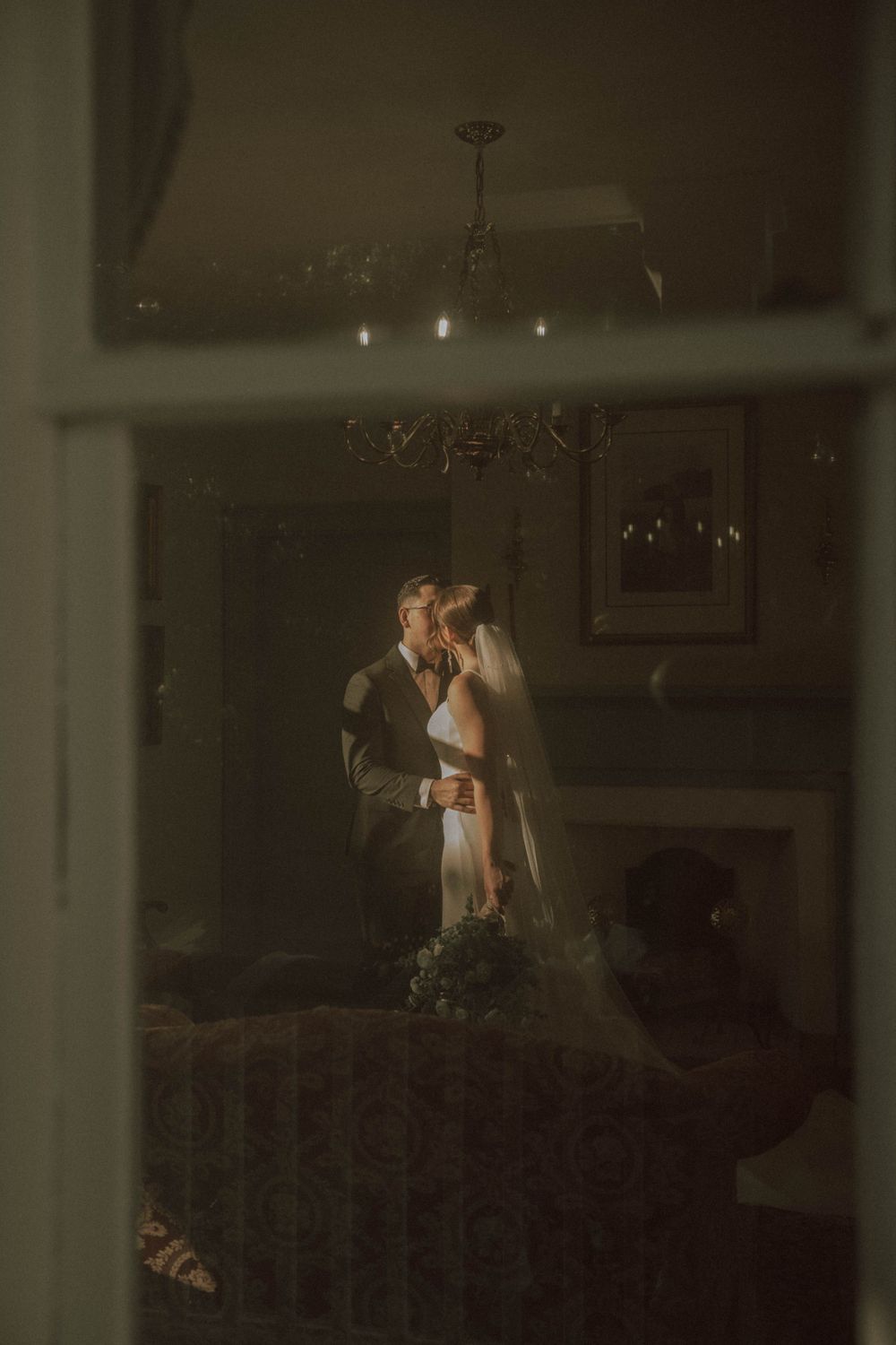 Artistic wedding photography shot through window panes showing couple in dramatic lighting.