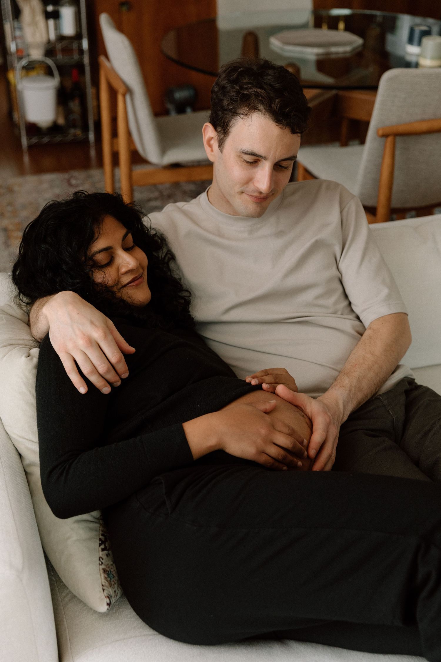 A couple relaxes together on a cozy white couch in a warmly lit living room.