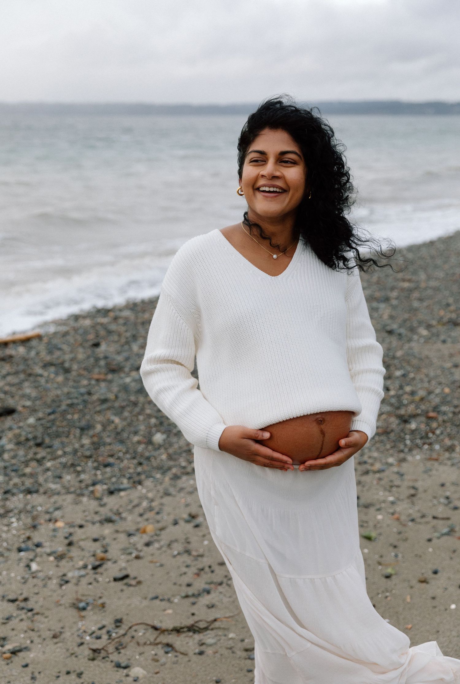 Expectant mother in white sweater and dress smiles joyfully while standing on a rocky beach with ocean in background.
