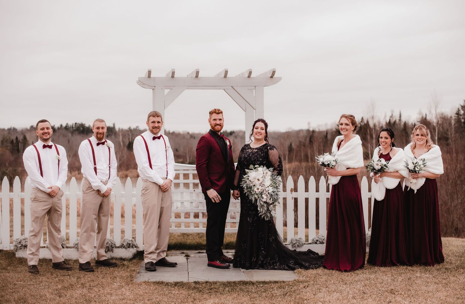 Wedding party members lined up against a white picket fence in formal attire.