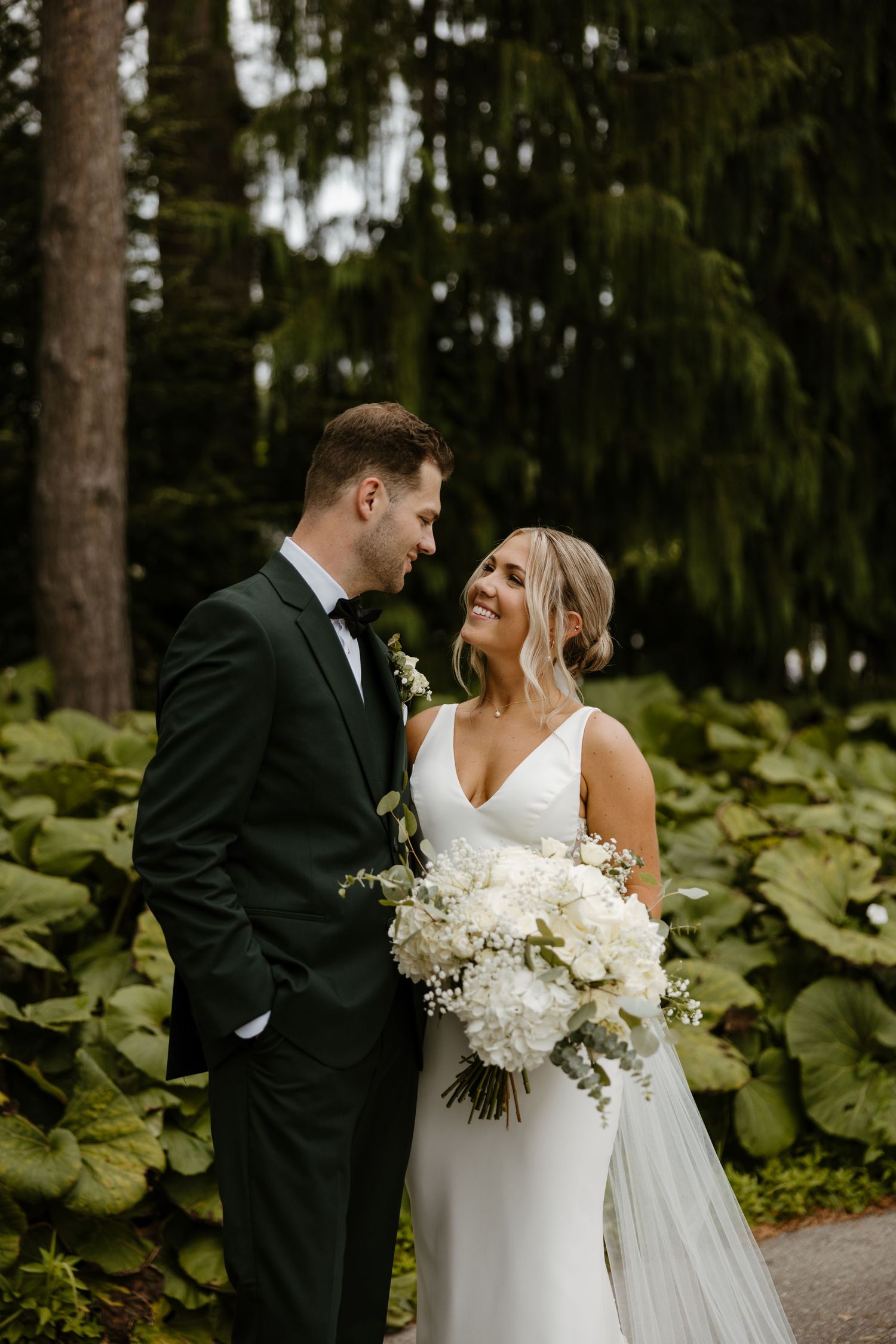 A bride and groom stand together in a lush, green garden setting, the bride holding a bouquet of white flowers. Fort Worth Texas.
