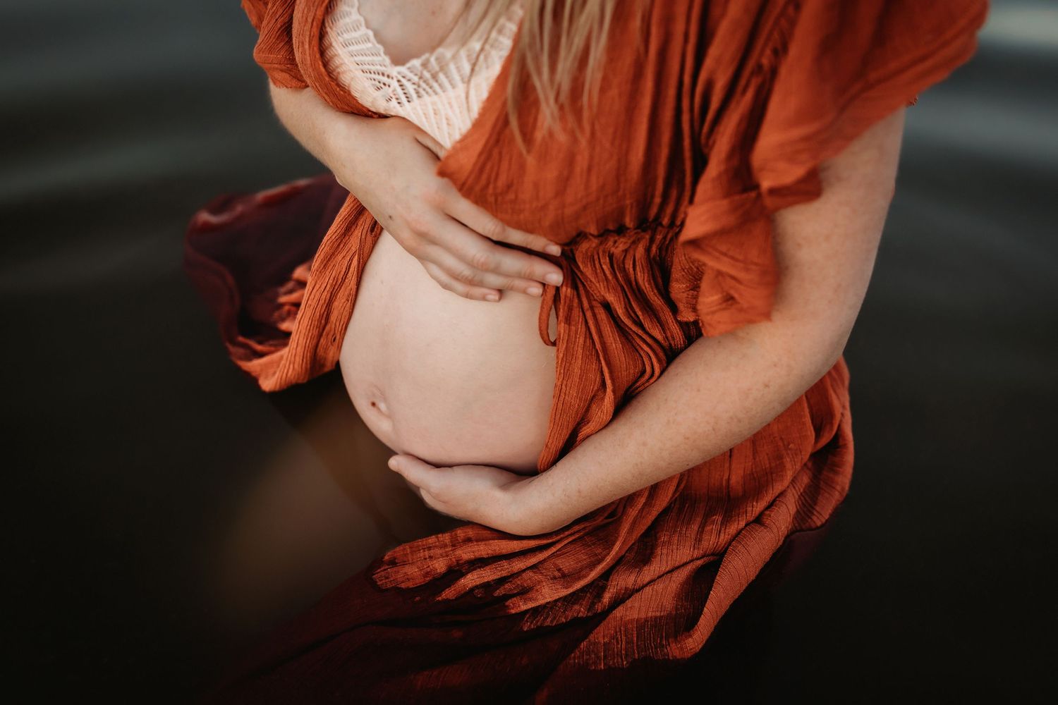 A pregnant woman in a flowing rust-colored dress poses in the water during a maternity photoshoot.
