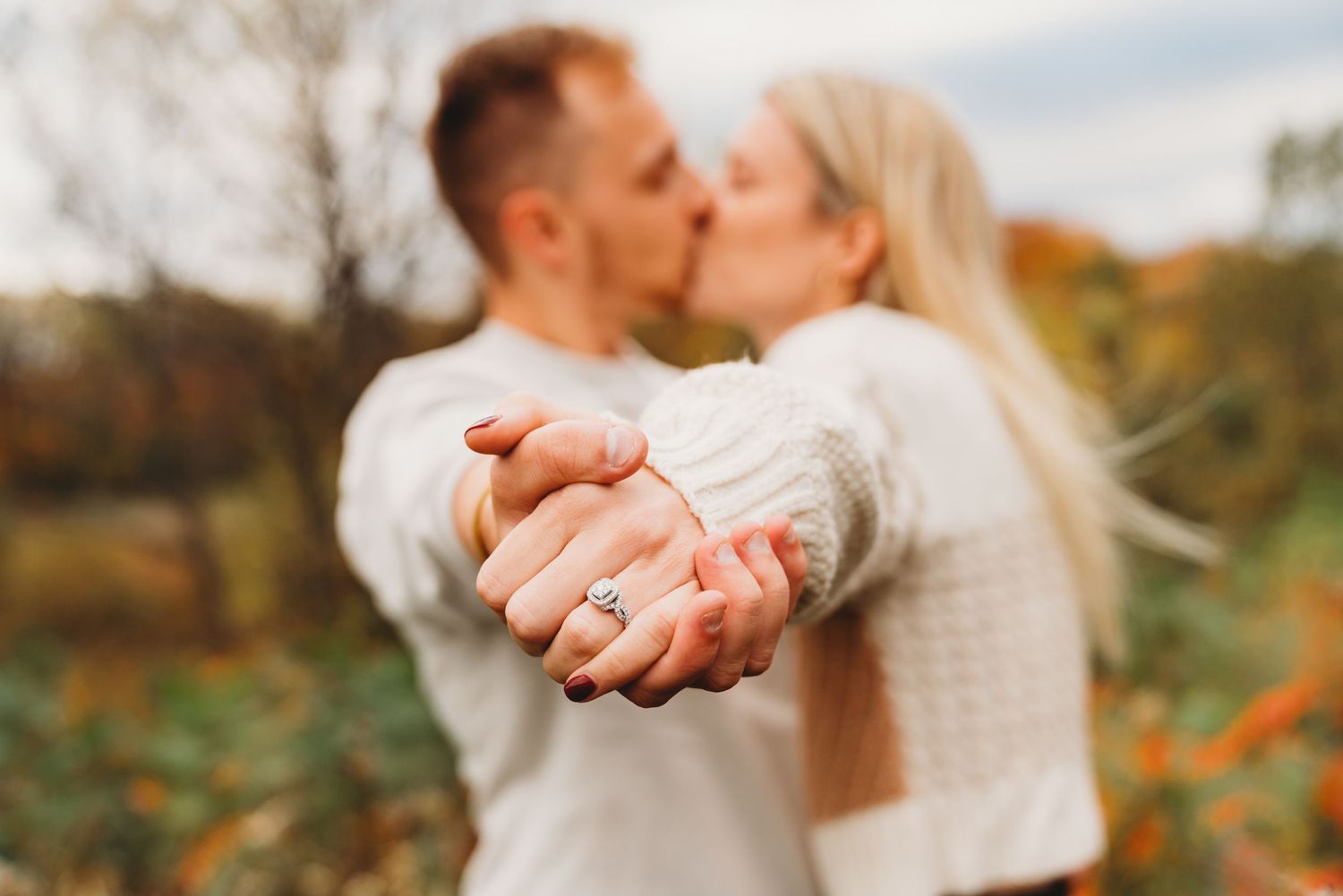 Close-up of engaged couple's clasped hands while sharing a romantic autumn kiss.