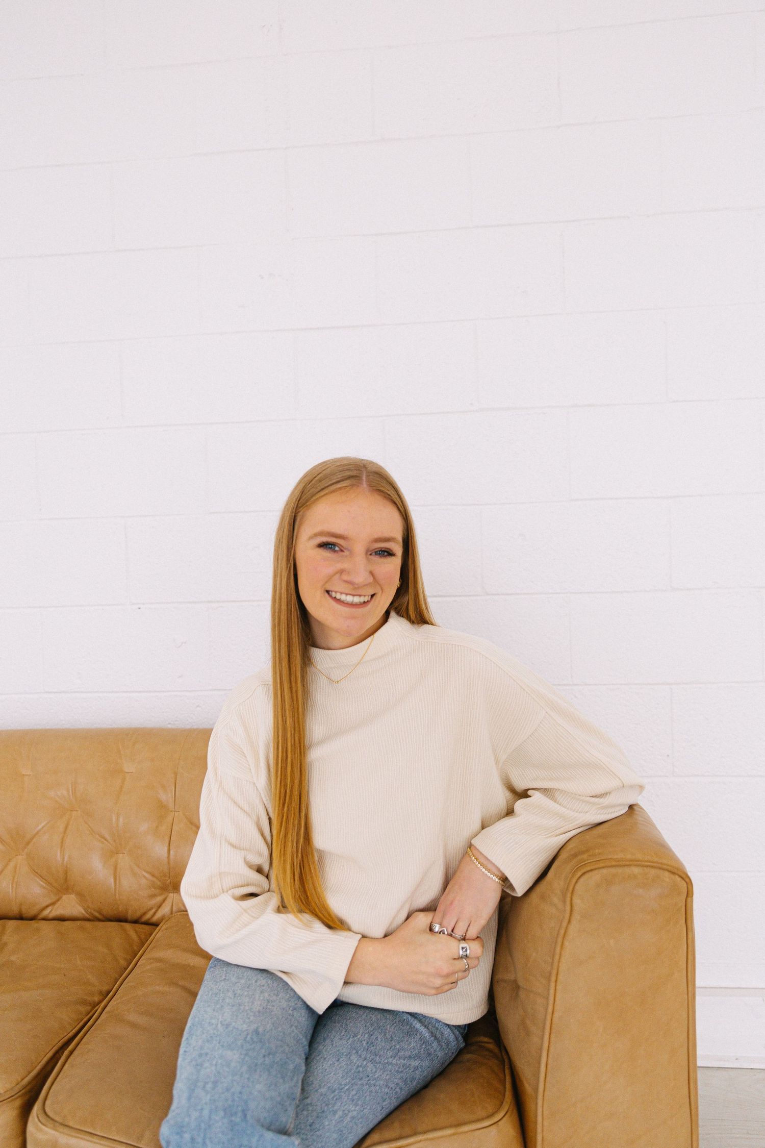 Person in cream turtleneck relaxes on brown leather couch during casual photoshoot.