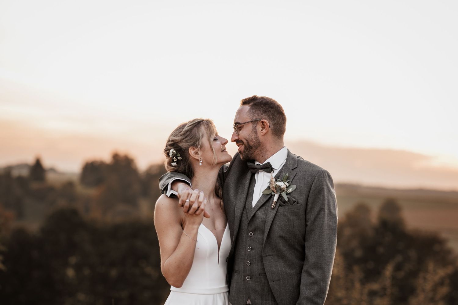 Ein romantisches Hochzeitsporträt bei Sonnenuntergang zeigt ein Paar, das sich auf einem Hügel umarmt, mit Bergen im Hintergrund