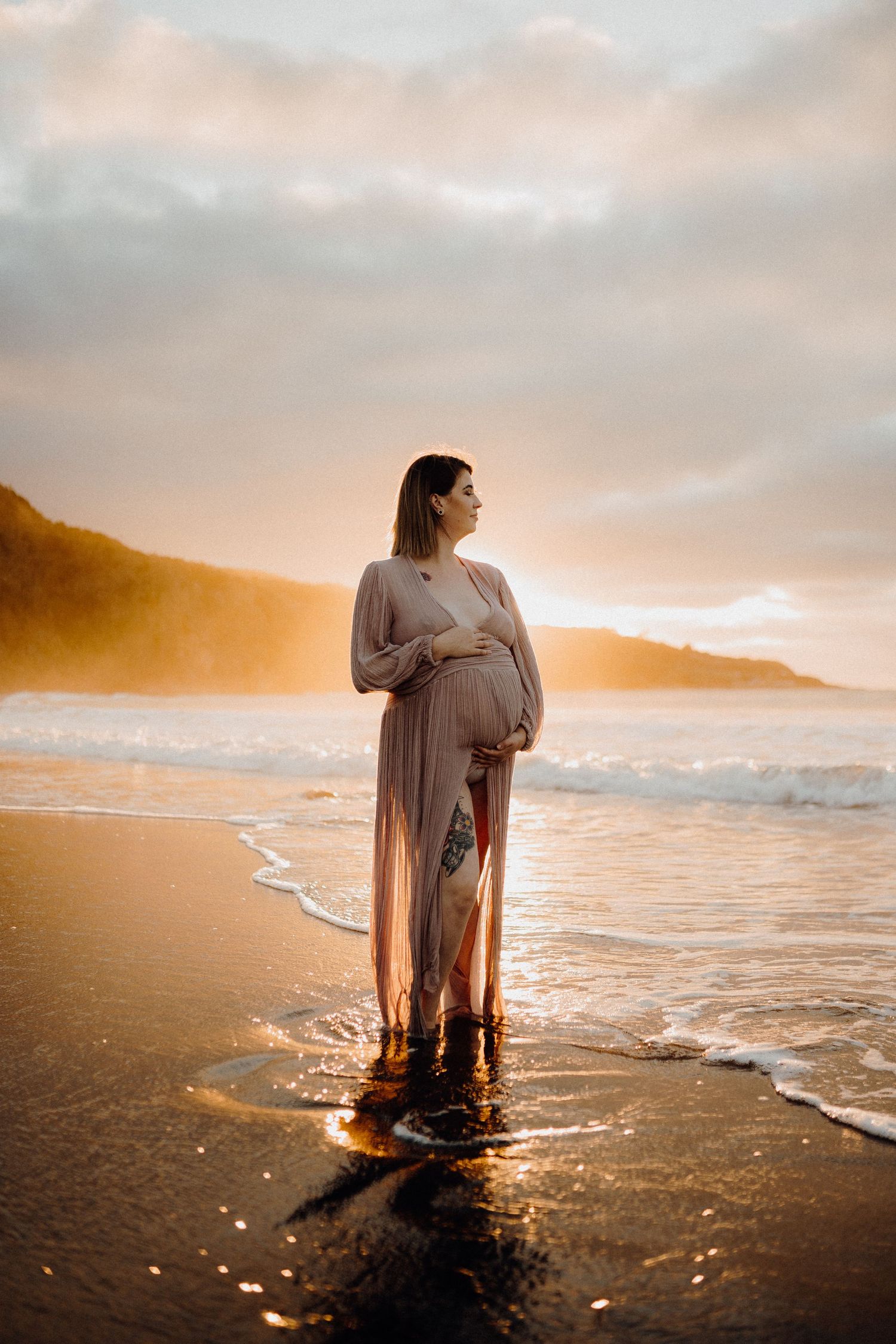 A silhouetted figure in a flowing dress stands in ocean waves at sunset.