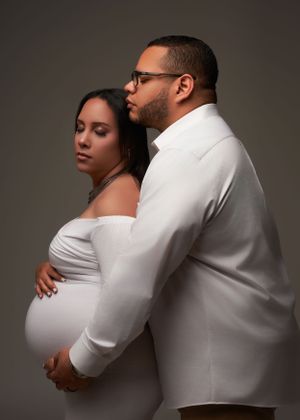 An intimate maternity portrait of a couple in white clothing against a dark background.