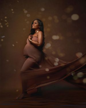 A maternity portrait against a brown backdrop with flowing fabric and soft bokeh lighting creating an ethereal glow.