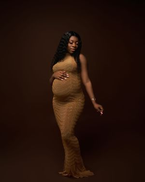 Expectant woman in a fitted gold gown poses elegantly against a dark brown studio backdrop.