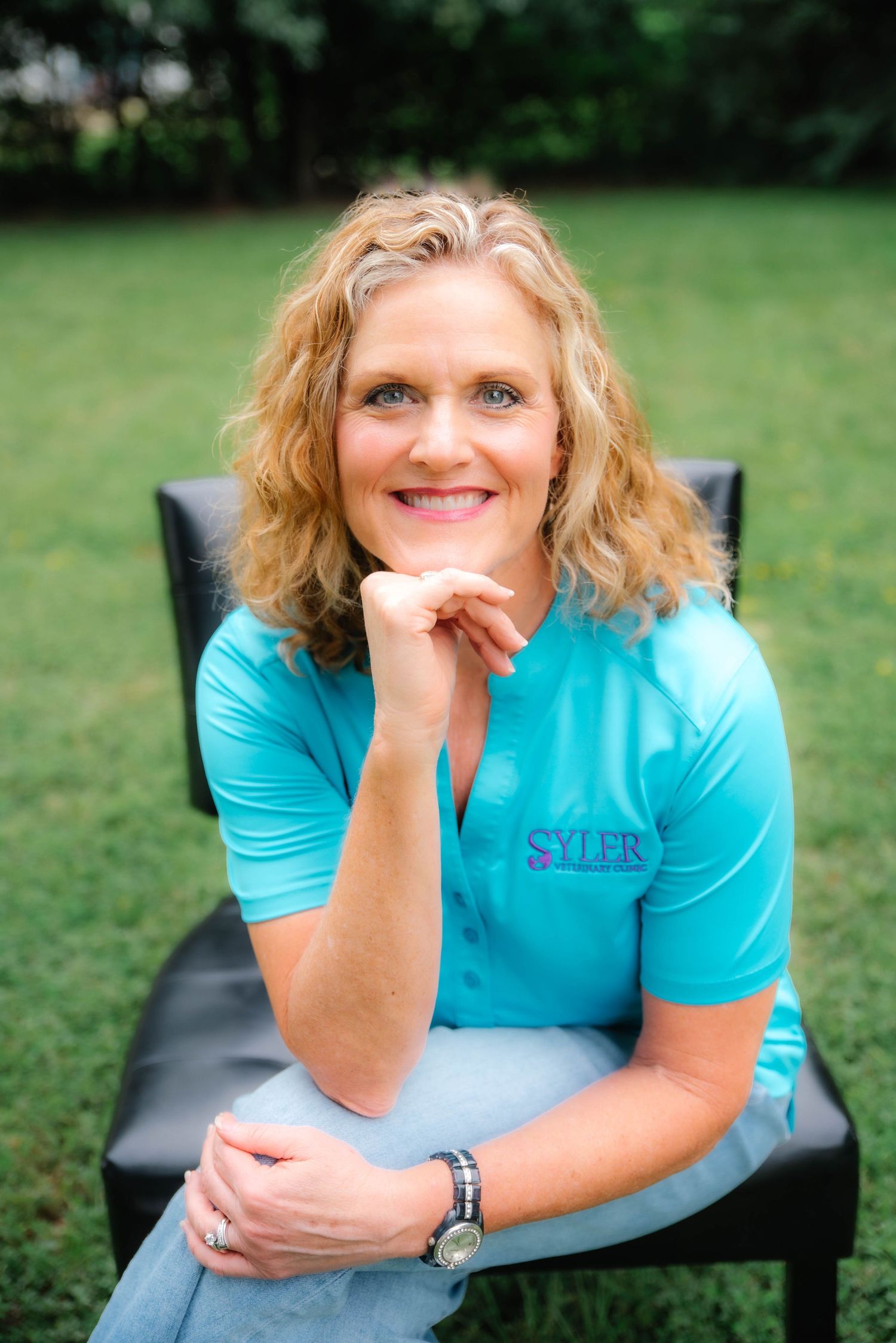 Smiling blonde woman in turquoise Syler polo shirt sitting outdoors on black chair with green grass background.