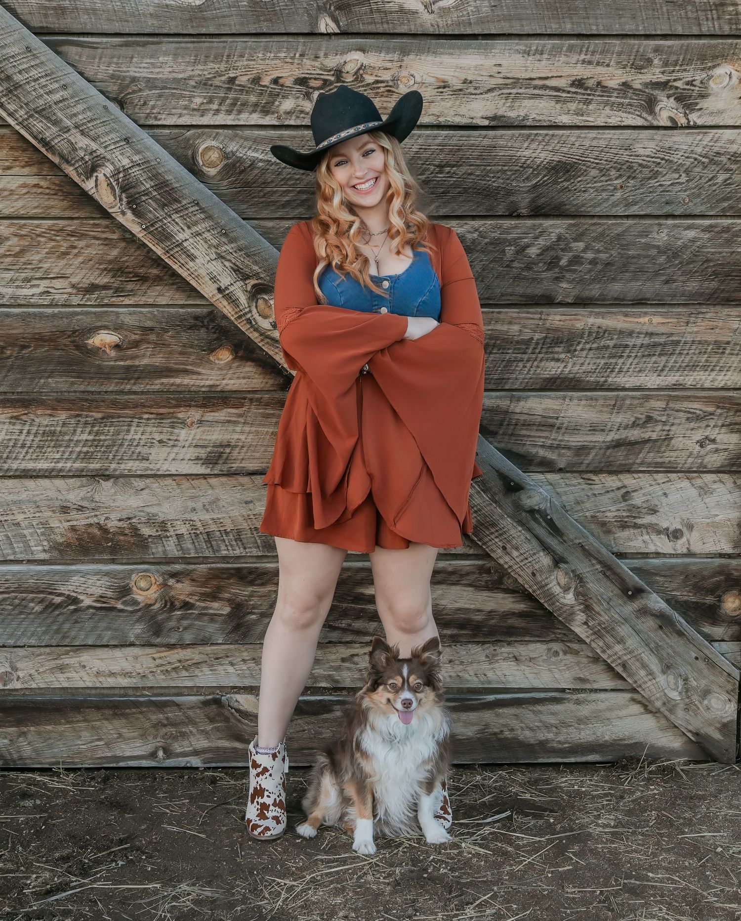 A woman in a rust-colored dress poses with a small fluffy Shih Tzu dog against a rustic wooden barn wall background.